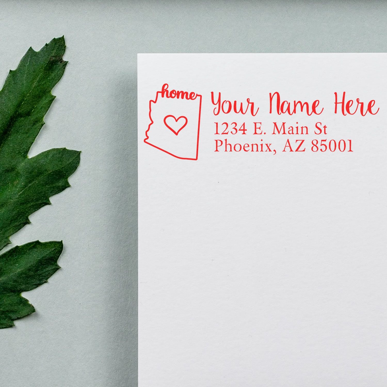 Wood Handle Arizona Custom Address Rubber Stamp on white paper with red ink, featuring a heart and state outline design, next to a green leaf on a light gray background.