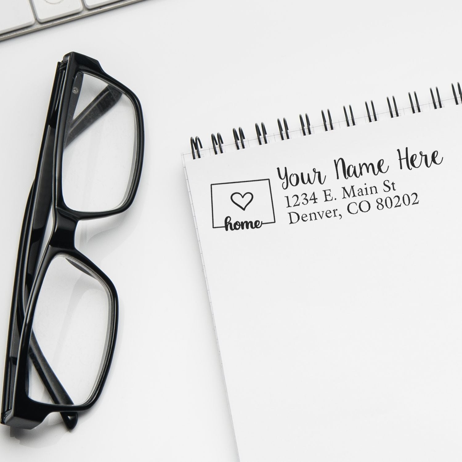 Glasses beside a notepad stamped with the Colorado Self-Inking State Love Address Stamp, featuring a heart and home design, displaying a sample address in Denver, CO.