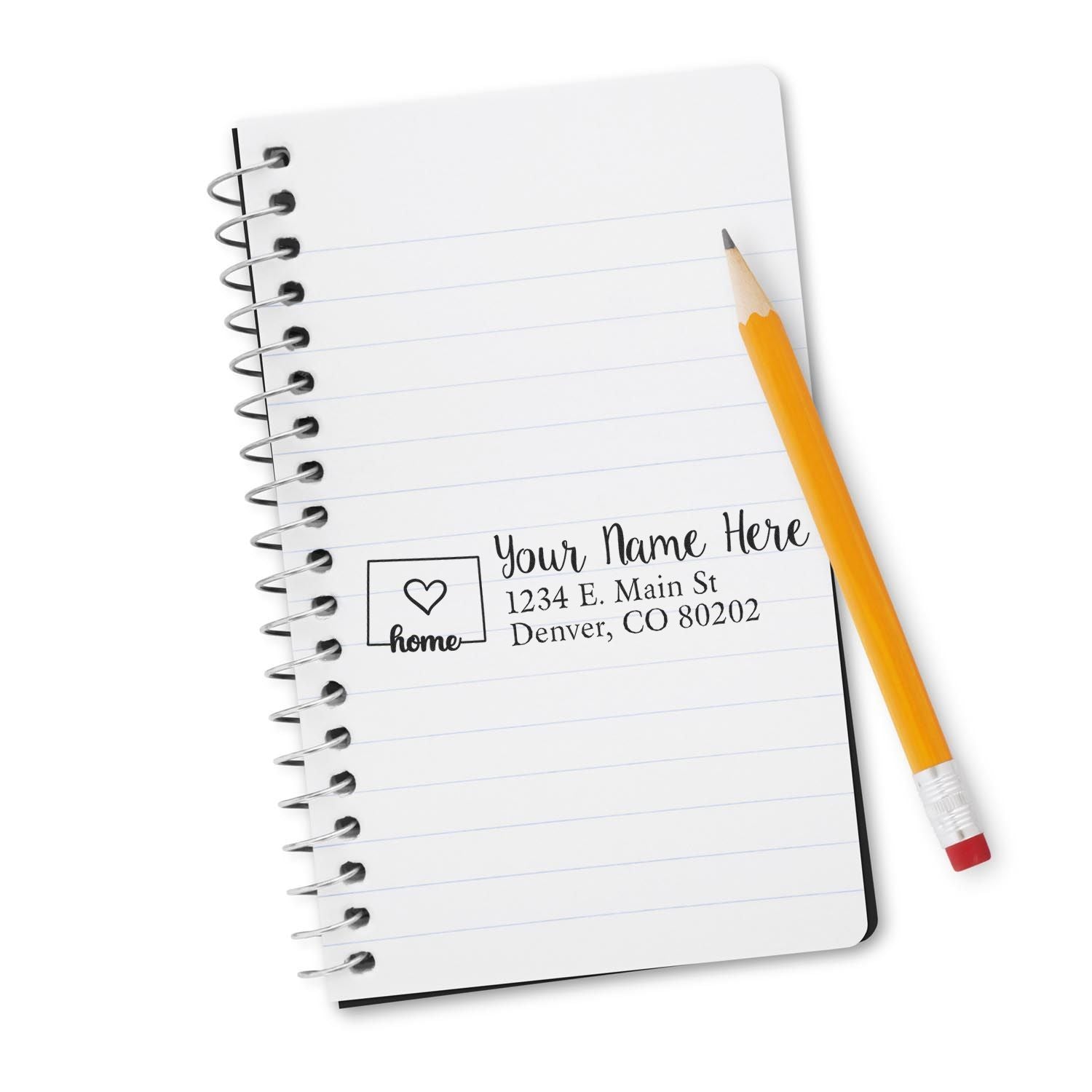 A Slim Pre-Inked Colorado State Love Custom Address Stamp is displayed on a spiral notebook page with a pencil beside it, showcasing a sample address in a stylish, personalized design.