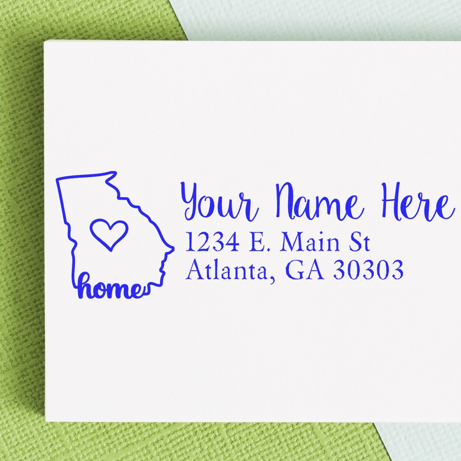 PSI Pre-Inked Personalized Georgia State Love Address Stamp on white envelope, featuring a blue outline of Georgia with a heart and home text, placed on a green textured surface.