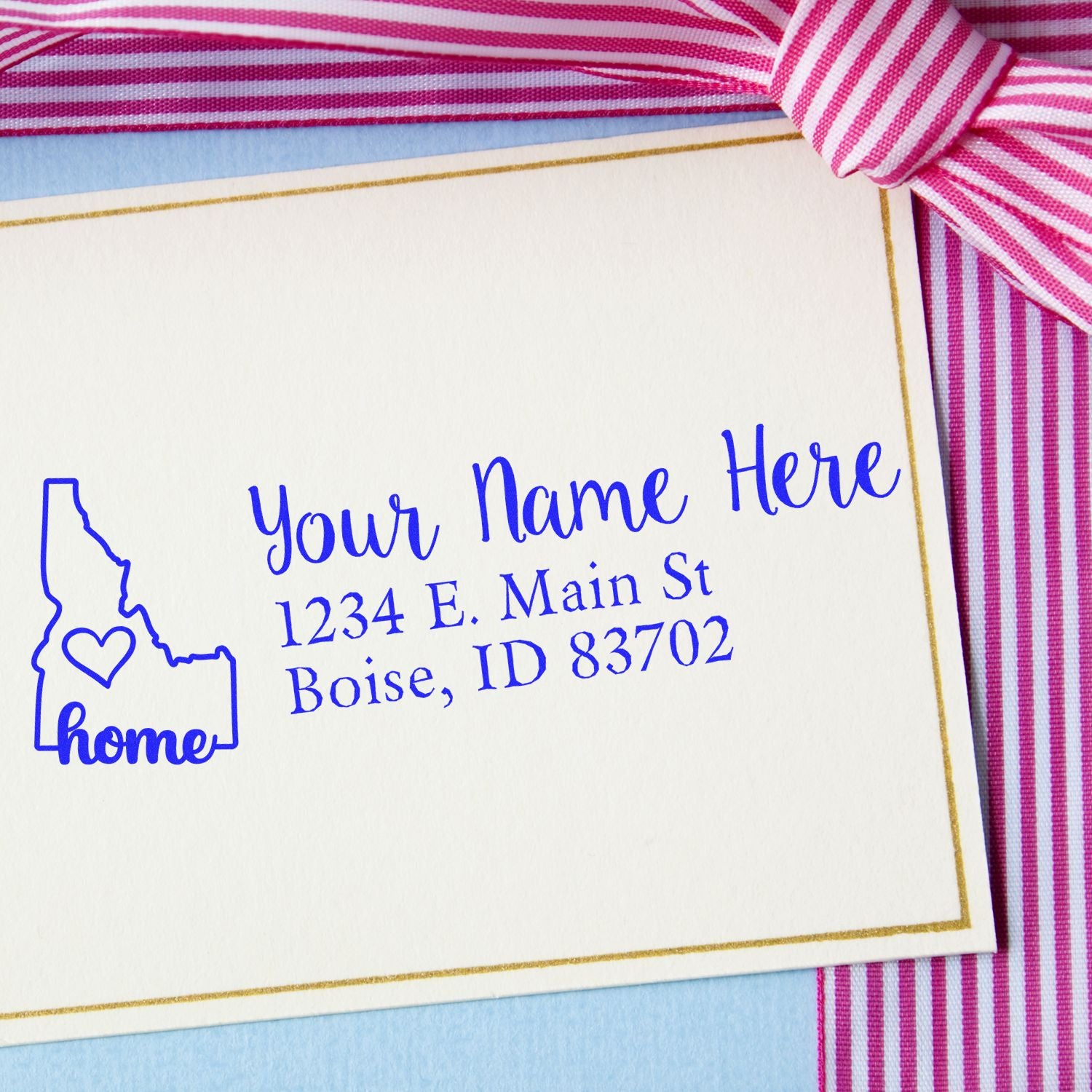 Idaho Self-Inking State Love Address Stamp on a card with a pink striped ribbon. Features a heart in Idaho outline, customizable name, and address in blue ink. Perfect for personalizing mail.