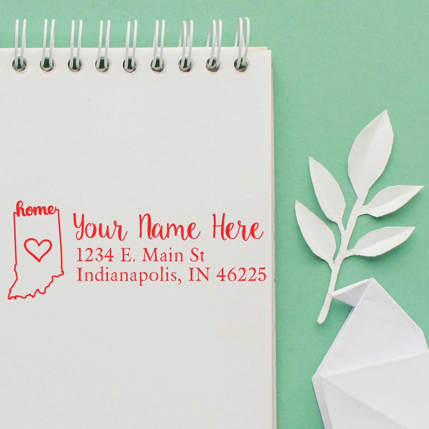 Wood Handle Indiana Custom Address Rubber Stamp on a notepad with a red imprint of an Indiana state outline, heart, and sample address. Green background with a paper leaf and envelope.