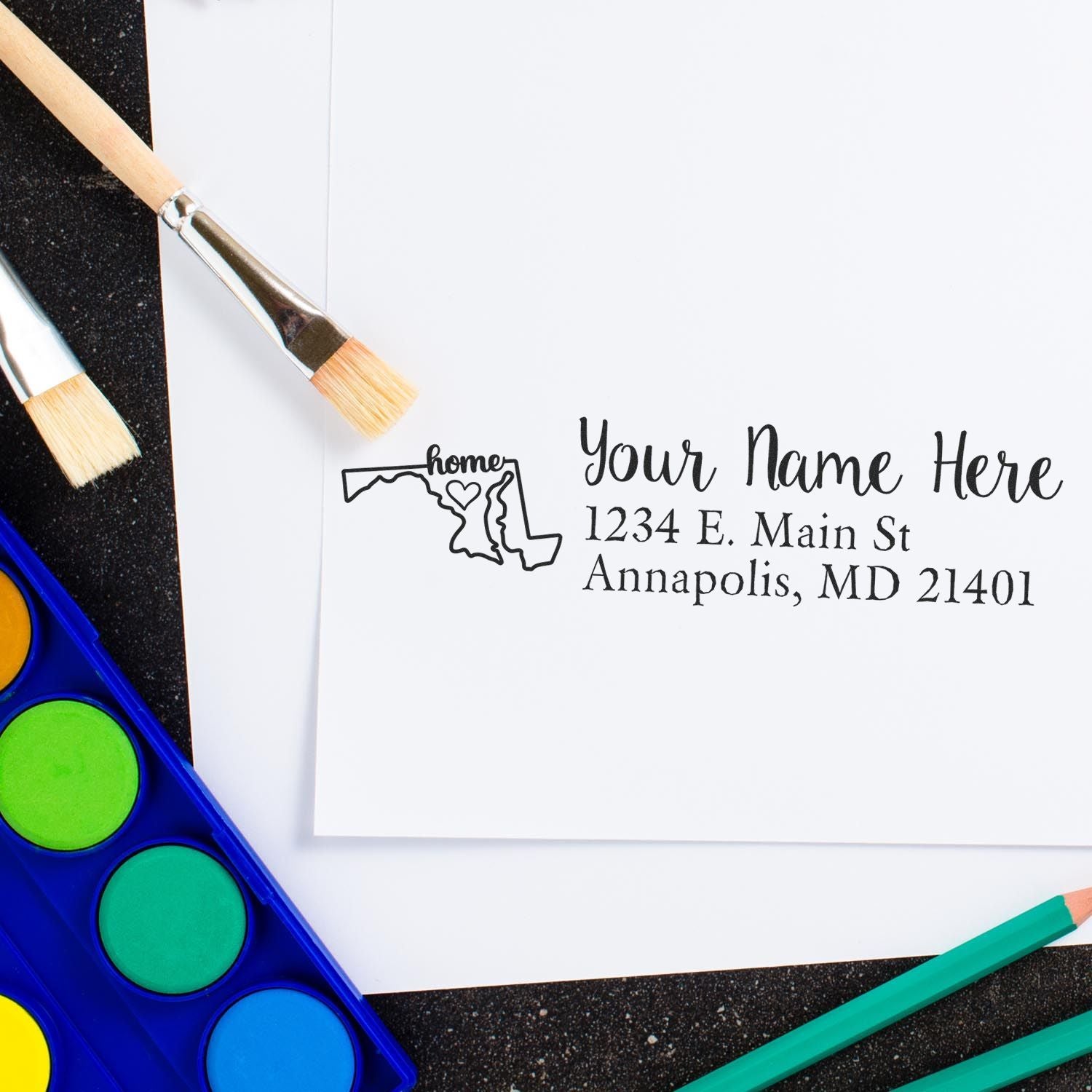 Wood Handle Maryland Custom Address Rubber Stamp on white paper, featuring a Maryland outline with home text. Surrounded by paintbrushes and watercolor paints on a black surface.