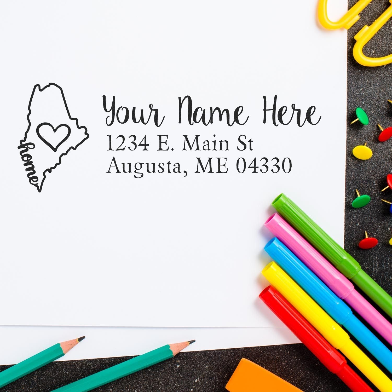 Slim Pre-Inked Maine State Love Custom Address Stamp on white paper with colorful pens and push pins. Features a heart inside Maine's outline, personalized with name and Augusta, ME address.