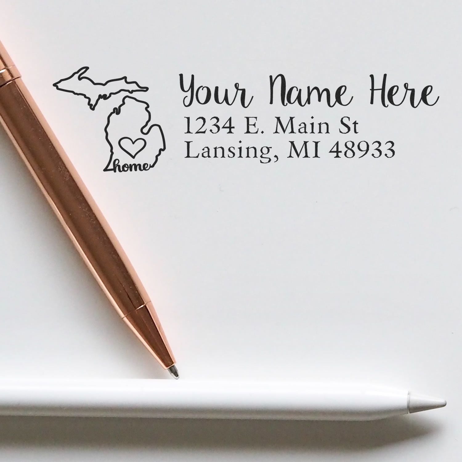 Slim Pre-Inked Michigan State Love Custom Address Stamp on white paper with a rose gold pen. Features a heart design within the Michigan state outline, personalized with name and address.