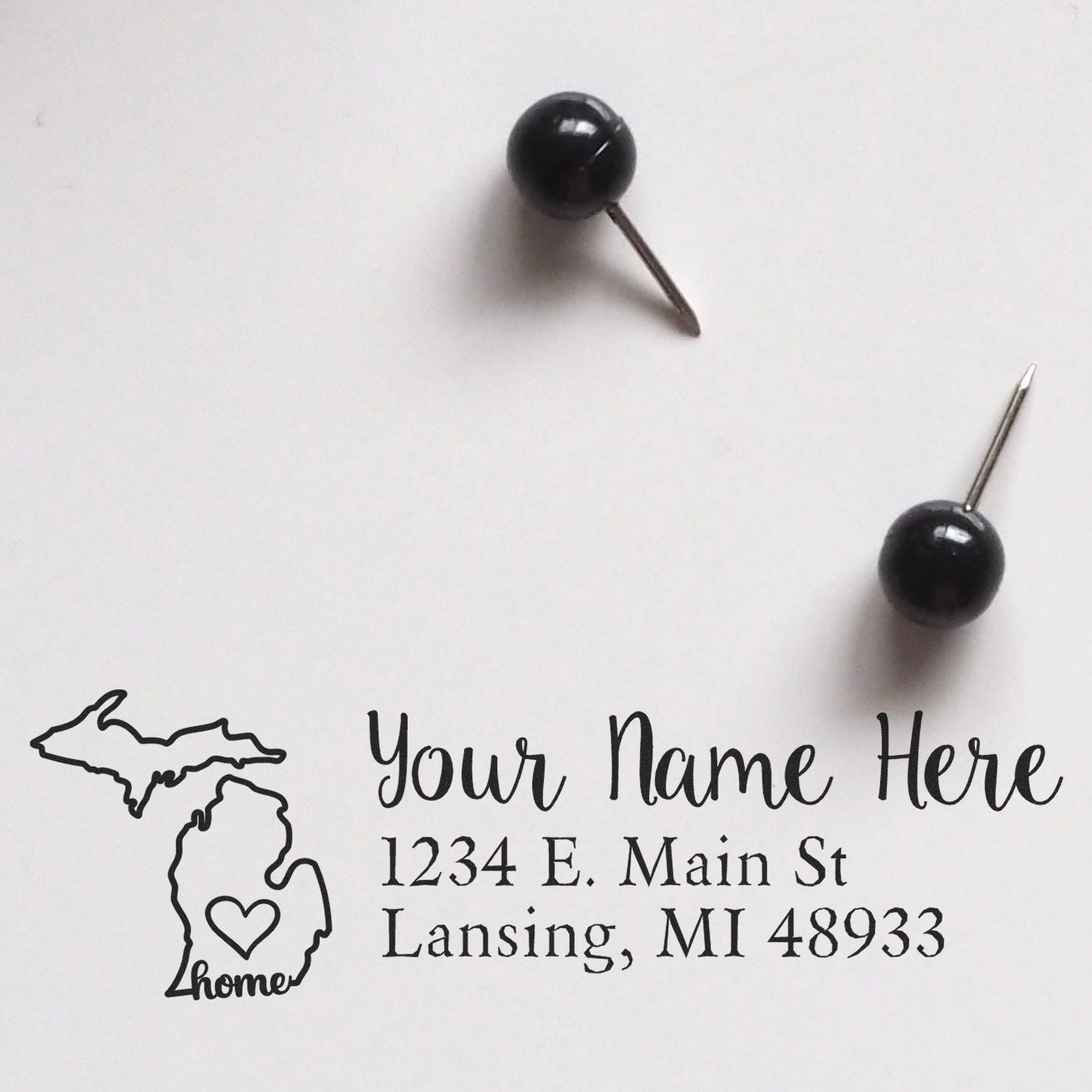 Slim Pre-Inked Michigan State Love Custom Address Stamp on white paper with two black push pins. Features a Michigan outline with a heart and customizable address text.