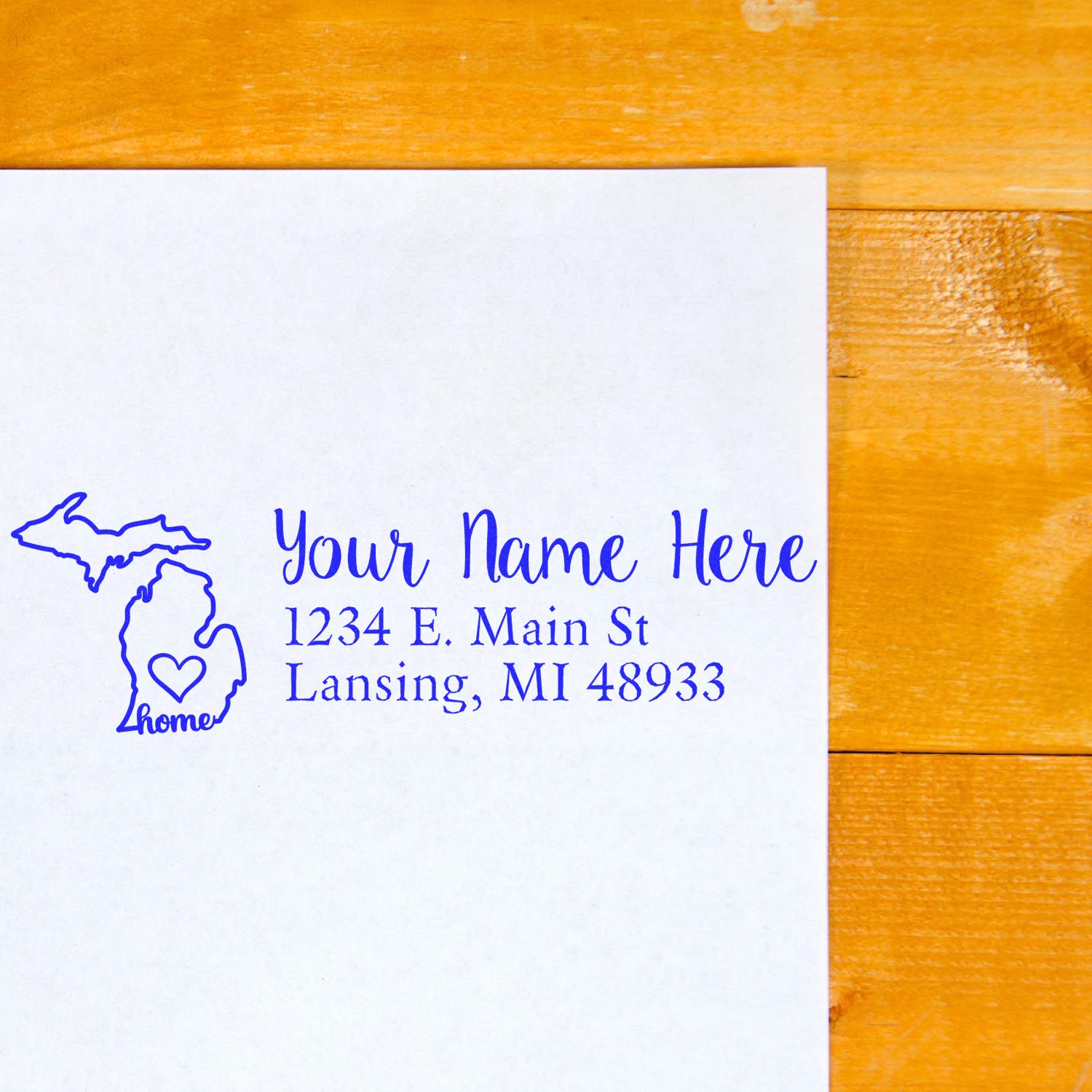 Wood Handle Michigan Custom Address Rubber Stamp on white paper with blue ink, featuring a Michigan outline and heart. Text reads Your Name Here, 1234 E. Main St, Lansing, MI 48933 on a wooden surface.