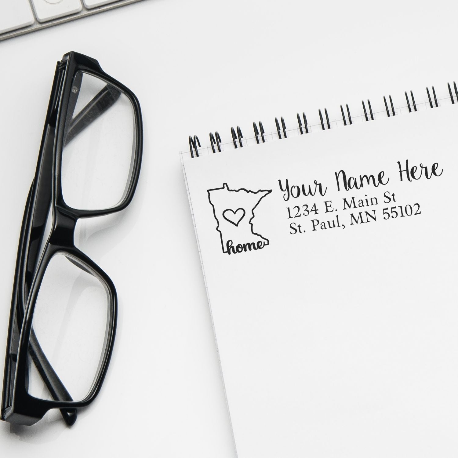 Black-framed glasses next to a notepad featuring the Minnesota Self-Inking State Love Address Stamp design, showing a heart inside the state outline with sample address text.