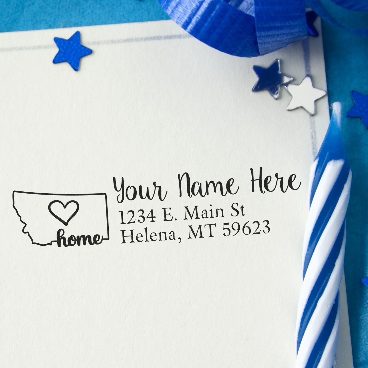 Montana Self-Inking State Love Address Stamp on an envelope with a heart design, surrounded by blue ribbon and stars, next to a blue and white striped candle.