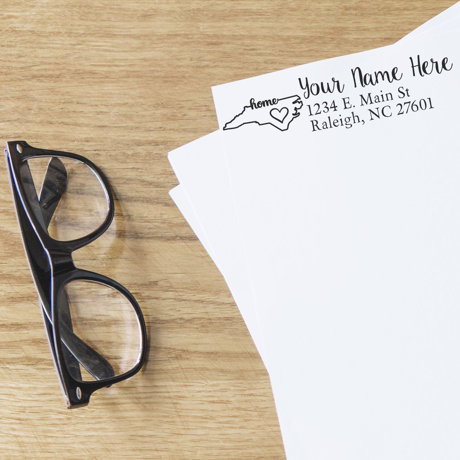 A pair of glasses rests on a wooden table beside papers stamped with the Wood Handle North Carolina Custom Address Rubber Stamp, featuring a heart and state outline design.