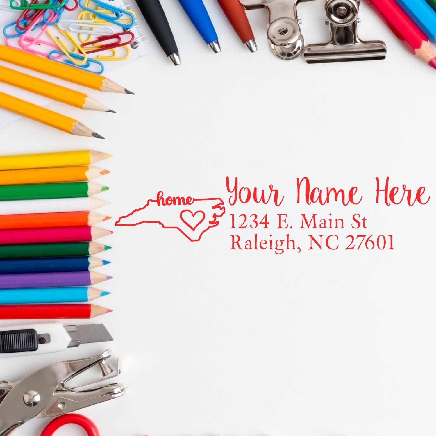 A PSI Pre-Inked Personalized North Carolina State Love Address Stamp on paper, surrounded by colorful pencils, paperclips, and office supplies. The stamp features a heart and the word home.