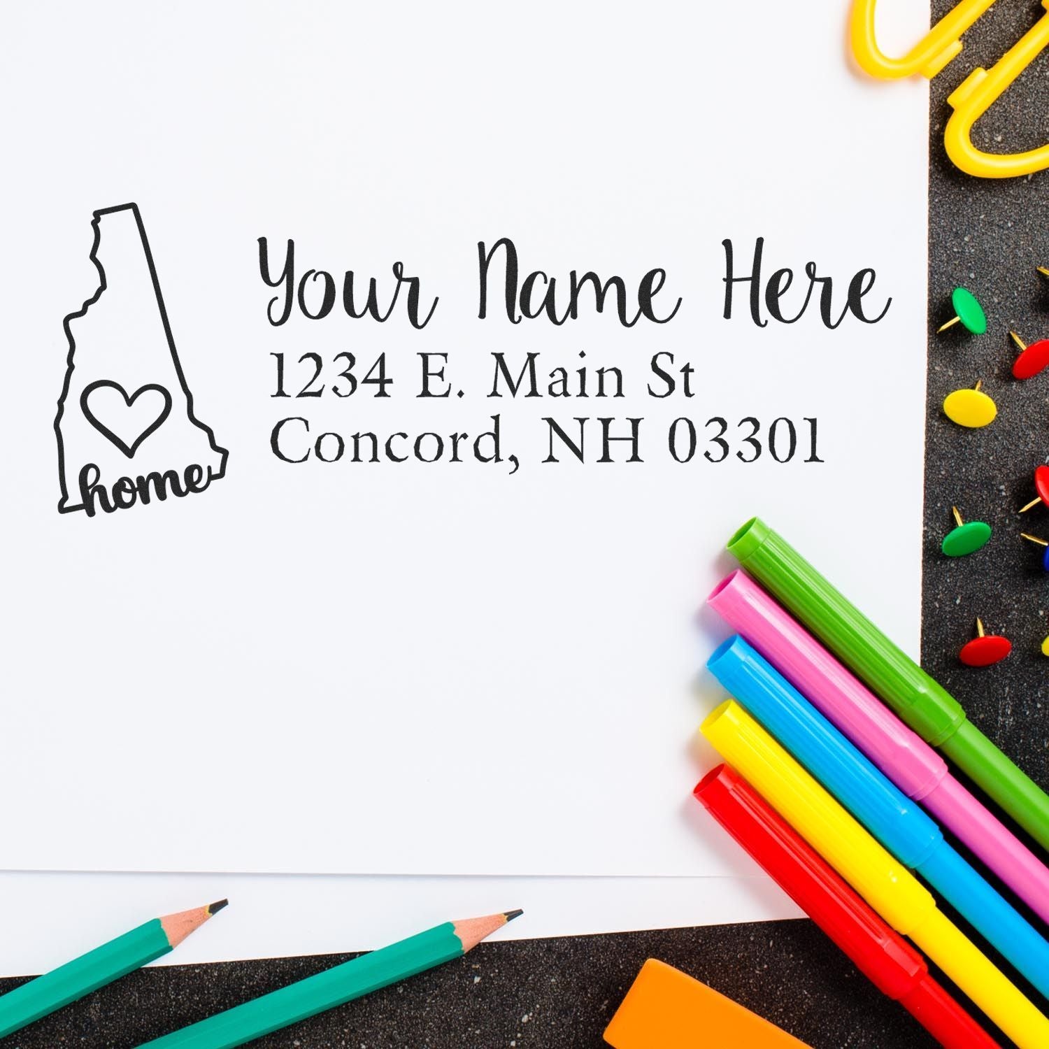 New Hampshire Self-Inking State Love Address Stamp on white paper with colorful pens and push pins. The stamp features a state outline with a heart and the word home, alongside customizable address text.