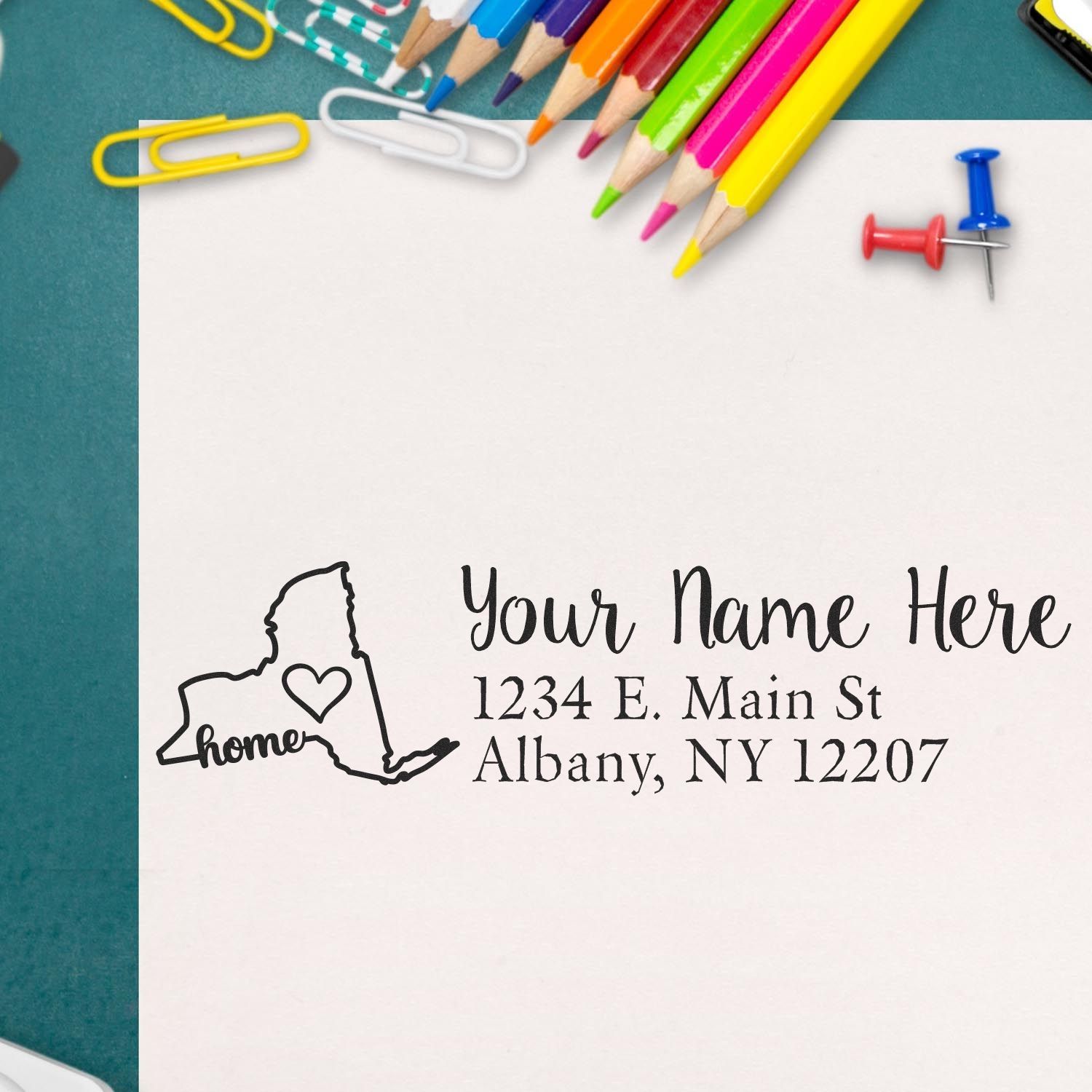 New York Self-Inking State Love Address Stamp on an envelope with colorful pencils and paperclips. Features a heart design within the state outline, personalized with name and address.