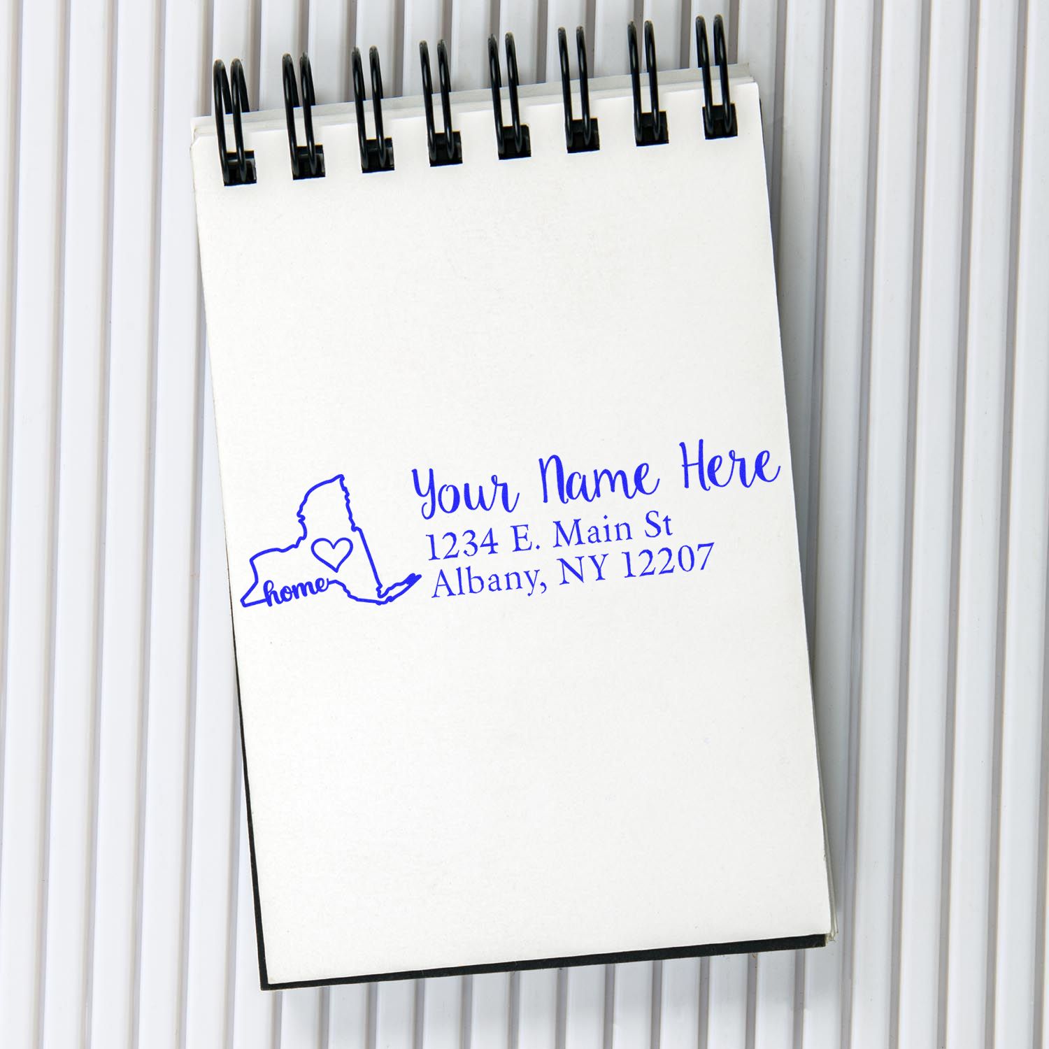 New York Self-Inking State Love Address Stamp on a notepad, featuring a blue outline of New York with home text and a heart, alongside customizable address fields.