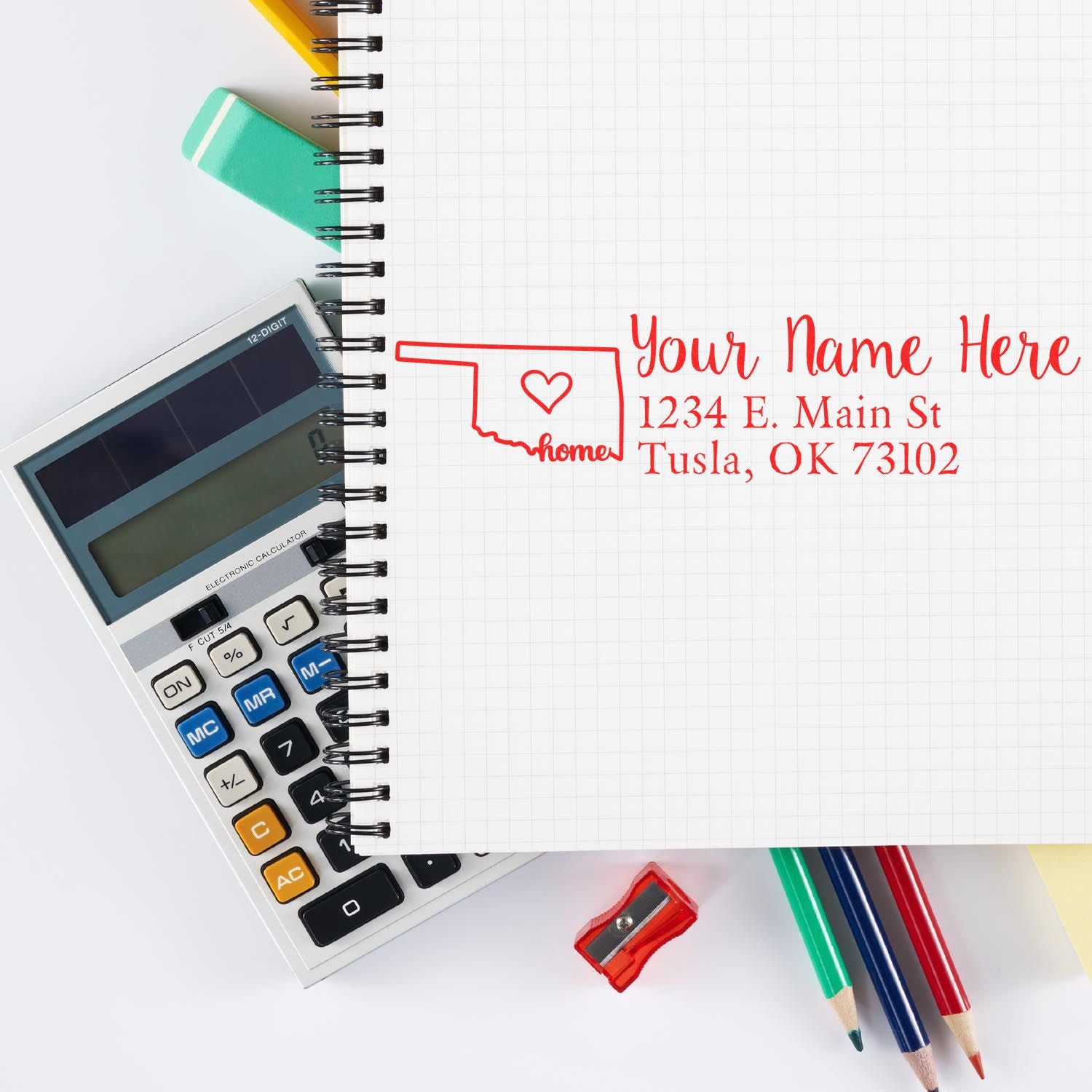 A notebook with the Oklahoma Self-Inking State Love Address Stamp imprint, featuring a heart and address, lies beside colorful pens, a calculator, and a sharpener on a white surface.