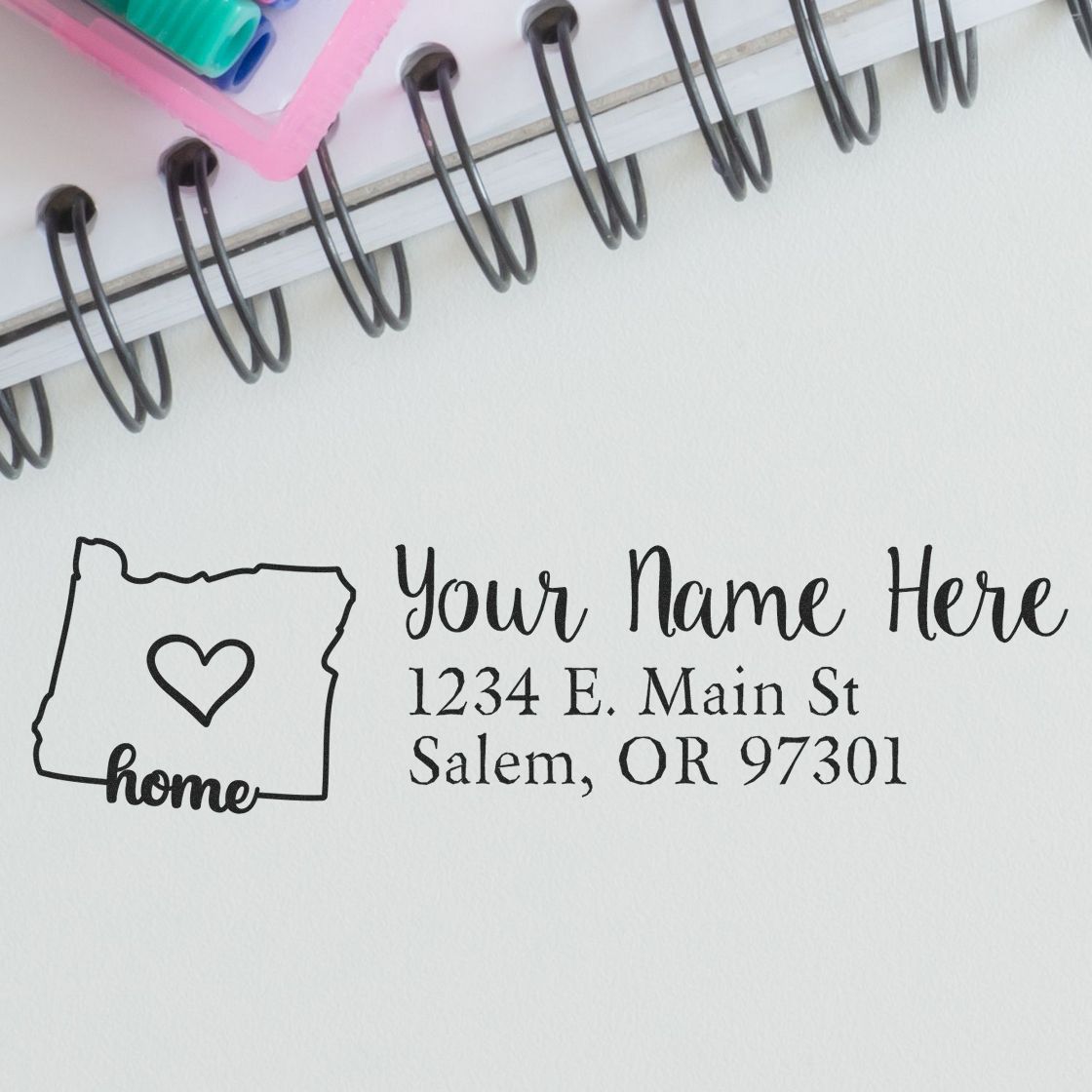 Oregon Self-Inking State Love Address Stamp on paper with a heart inside the state outline, personalized with Your Name Here and an address in Salem, OR. Nearby are a notebook and colorful pens.