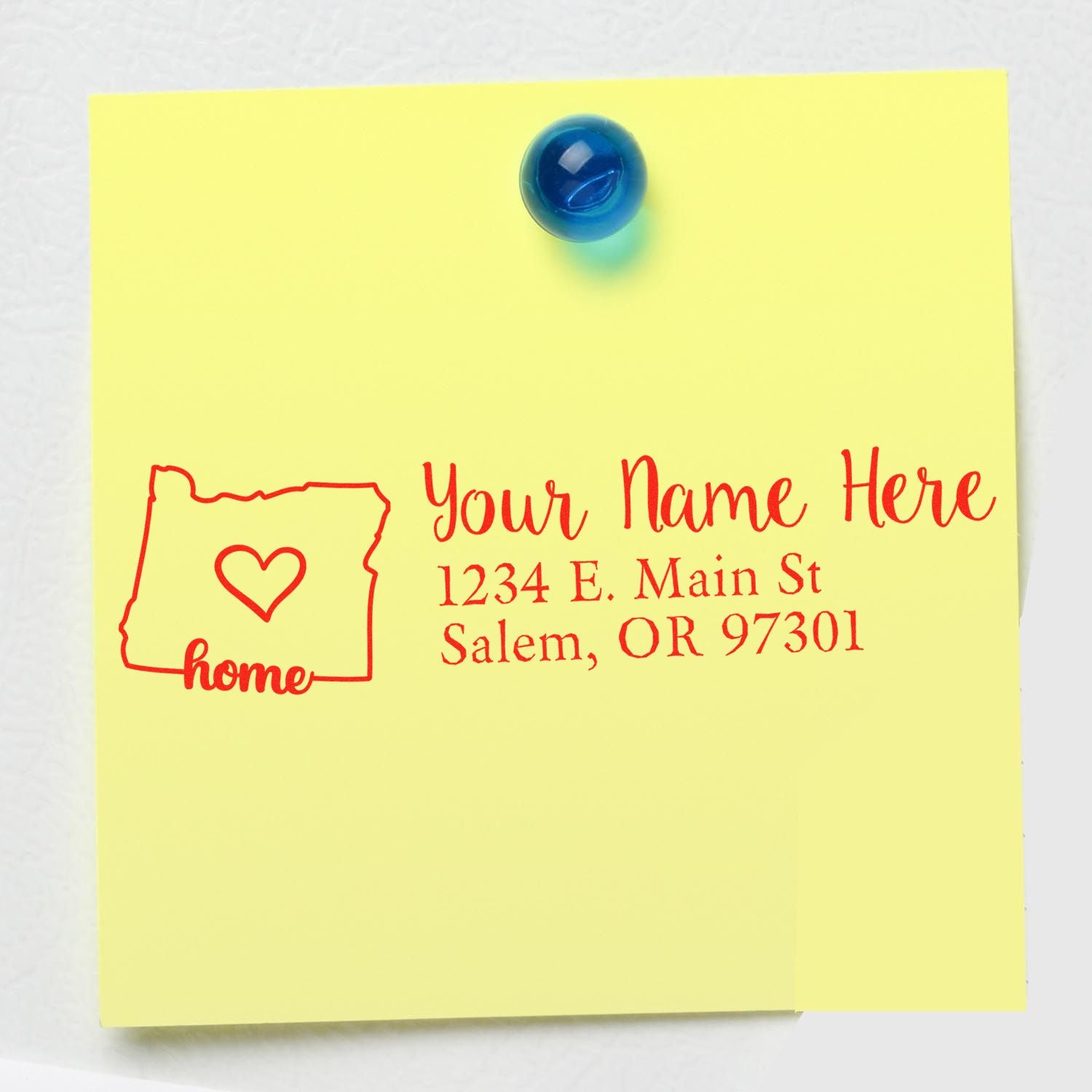 Yellow note with a red Slim Pre-Inked Oregon State Love Custom Address Stamp design, featuring a heart inside the state outline and sample address text. Blue magnet holds the note on a white surface.