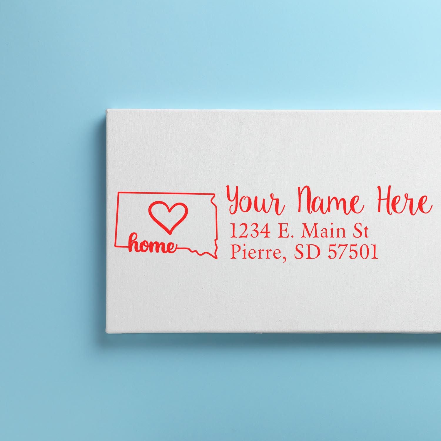 South Dakota Self-Inking State Love Address Stamp on a white envelope with red ink, featuring a heart inside the state outline and sample address text. Set against a light blue background.