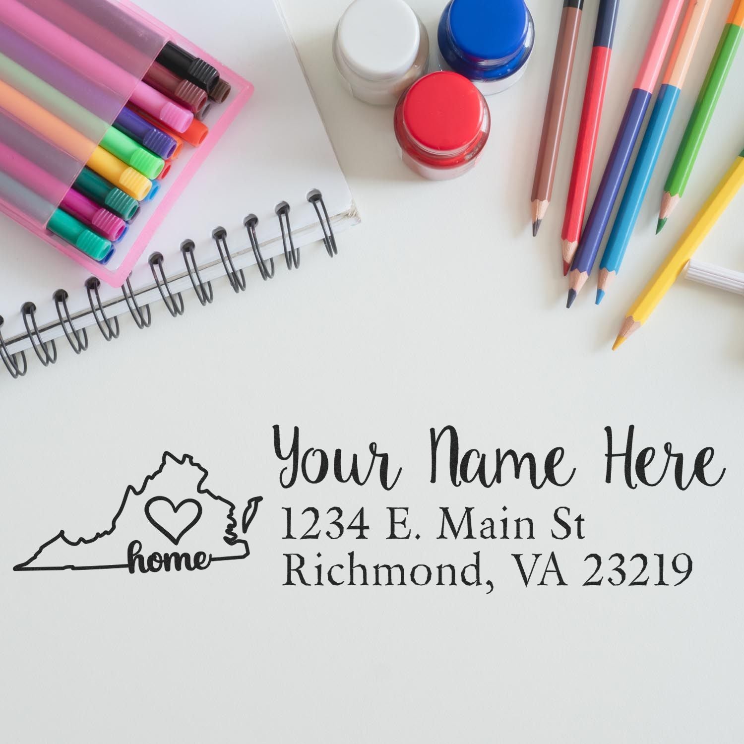 A PSI Pre-Inked Personalized Virginia State Love Address Stamp on paper, surrounded by colorful pens, markers, and a notebook, showcasing a custom address with a heart design in Virginia's outline.