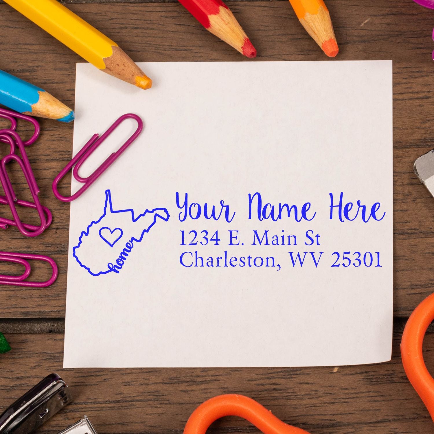 West Virginia Self-Inking State Love Address Stamp on a white paper with a blue imprint, surrounded by colorful pencils, paperclips, and a wooden desk background.