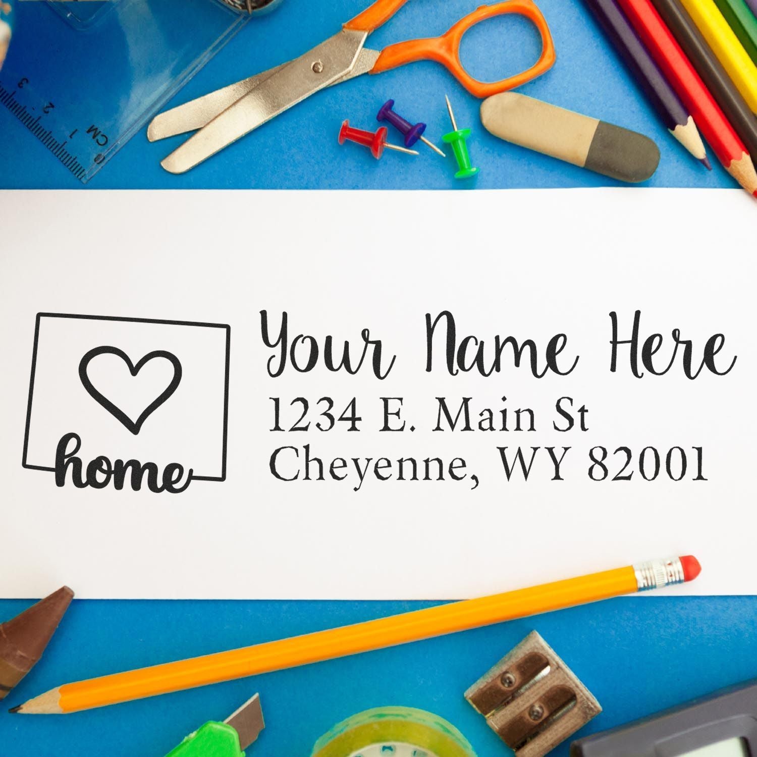 A PSI Pre-Inked Personalized Wyoming State Love Address Stamp on a white envelope, surrounded by colorful office supplies like scissors, pencils, and push pins on a blue background.