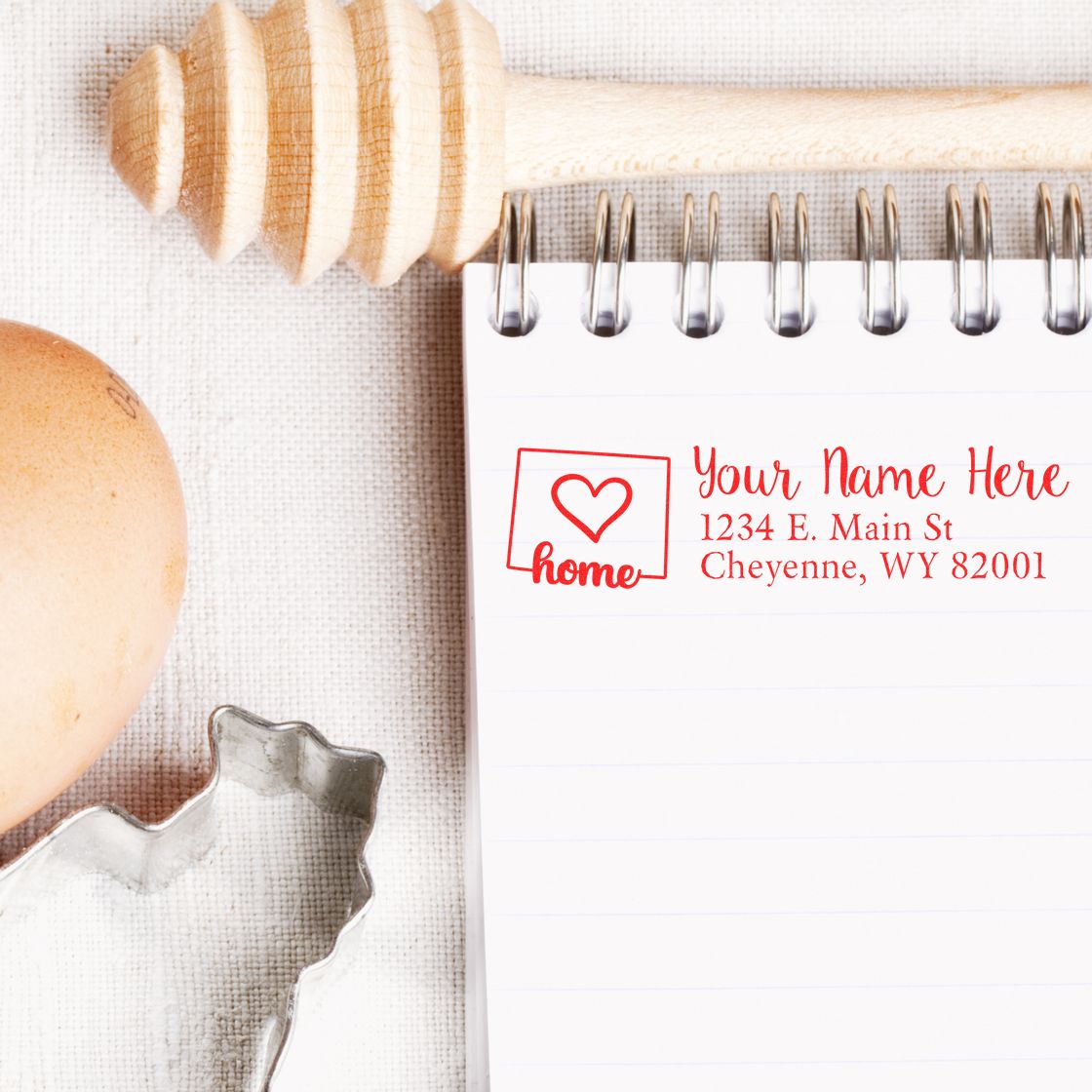 Wyoming Self-Inking State Love Address Stamp on a notepad with a heart design, next to a honey dipper, egg, and cookie cutter, showcasing a personalized address in red ink.