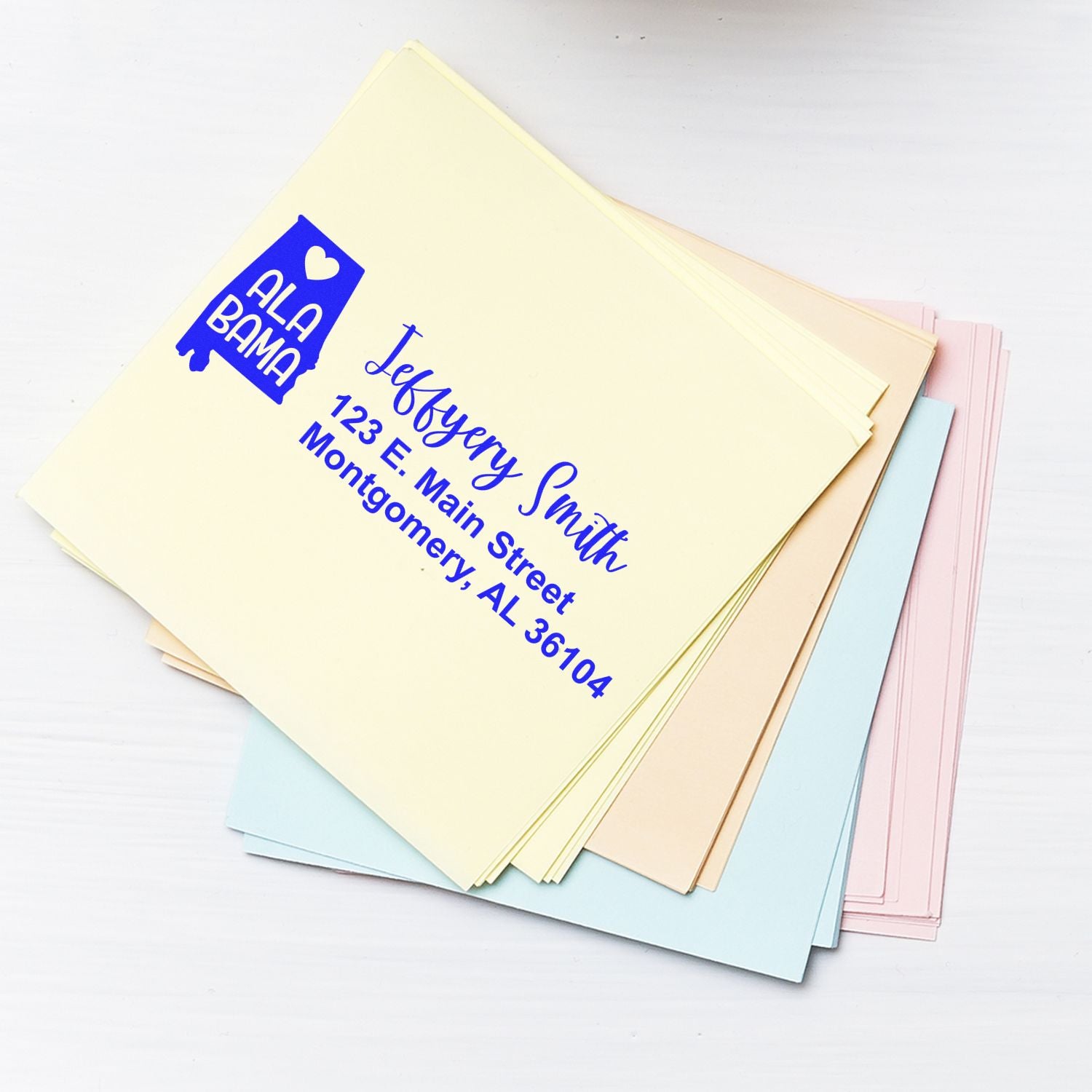 A stack of pastel envelopes featuring the Alabama State Love Personalized Address Stamp with a heart design, displaying the name and address in blue ink.