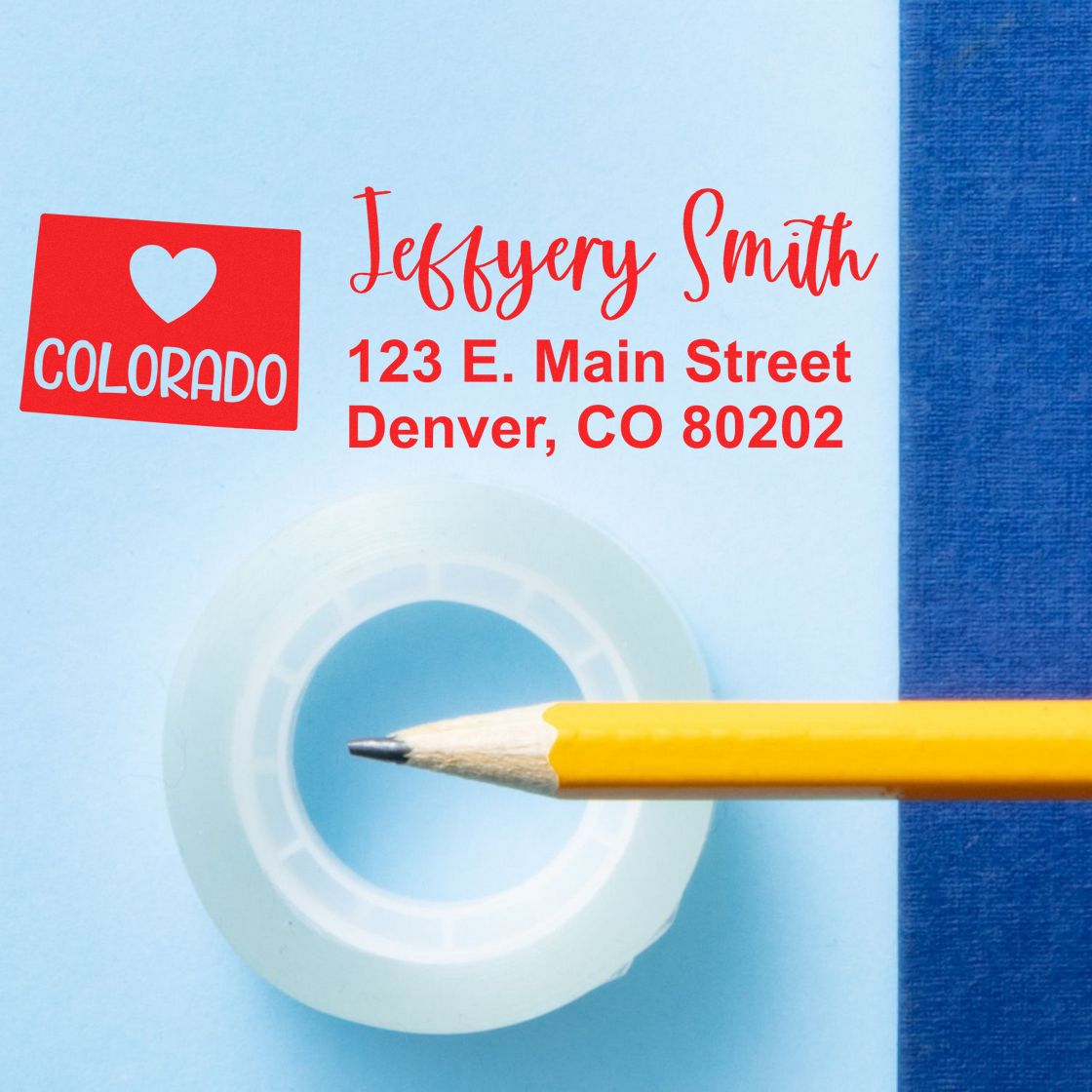 Image of the State Love of Colorado Custom Address Stamp Self-Inking, featuring a red heart design with Colorado text, next to a pencil and tape on a blue background.