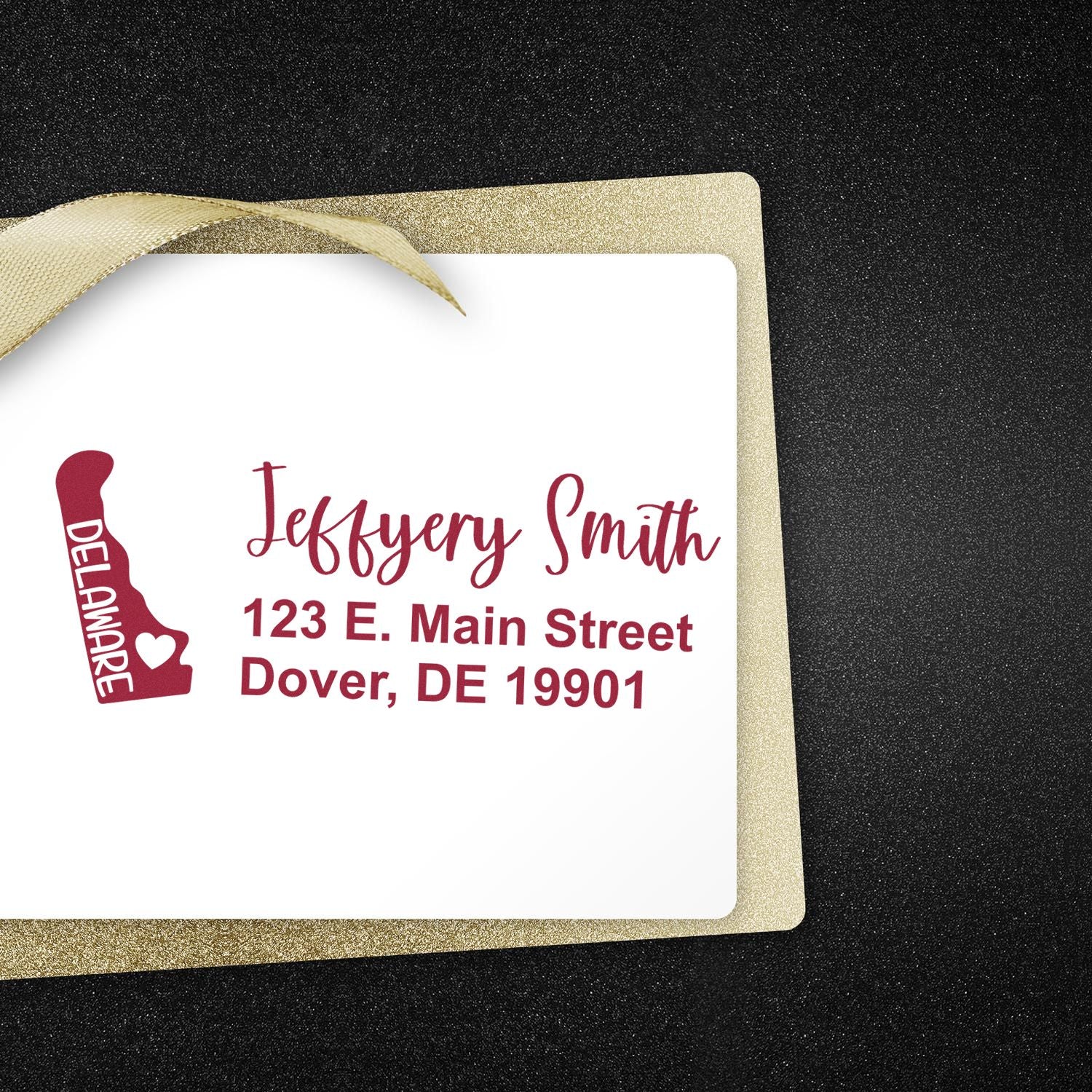 Slim Delaware Custom Address Stamp for Envelopes displayed on a white card with a gold ribbon. The stamp features a Delaware state outline and personalized address in red text.