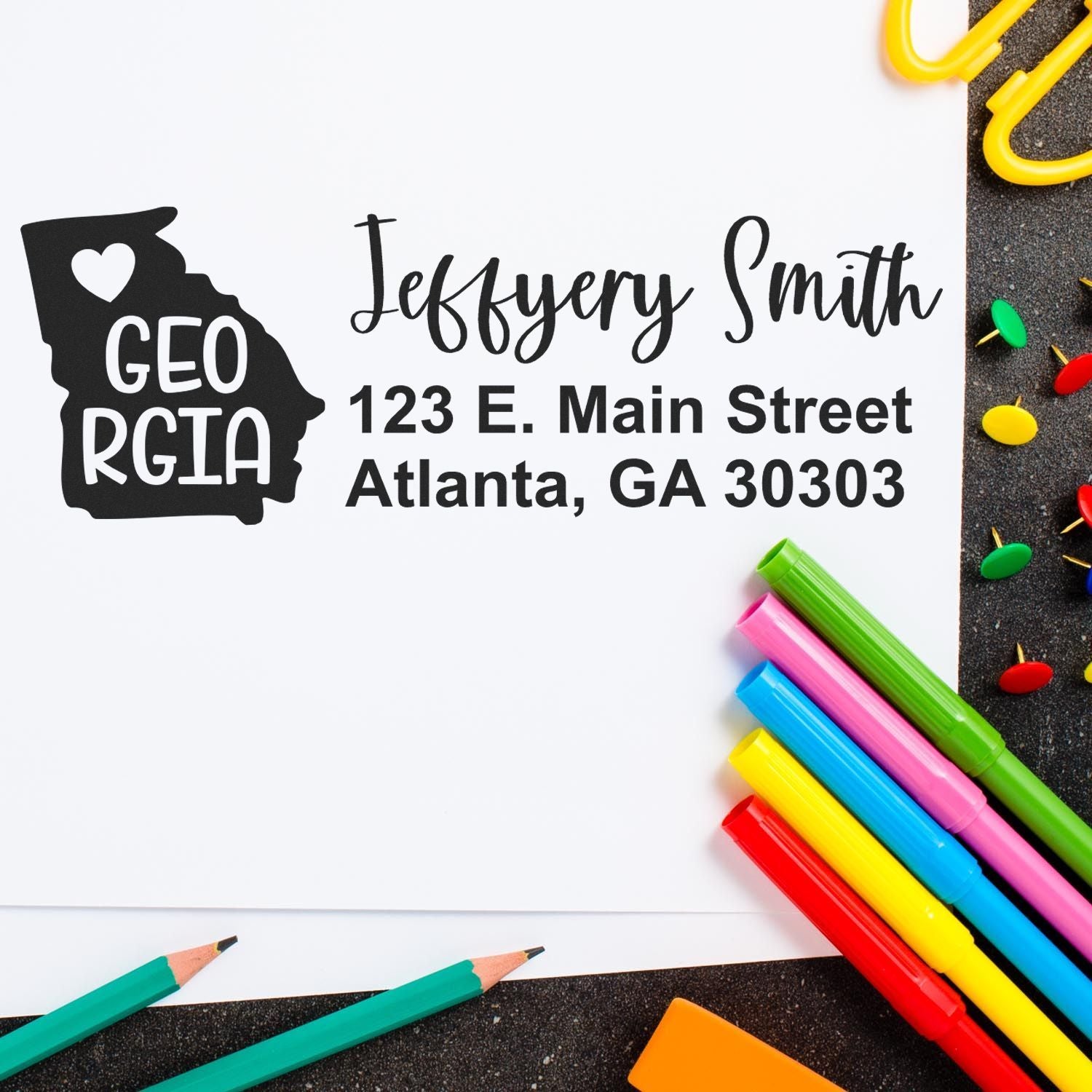 Image of the Georgia State Love Personalized Address Stamp on an envelope, featuring a heart and state outline. Surrounded by colorful pens and paperclips on a dark surface.