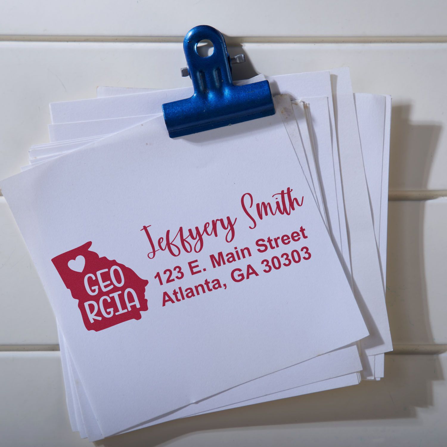 Stack of envelopes with a Georgia State Love Personalized Address Stamp featuring a red Georgia outline, heart, and custom address in red text. Blue clip holds the stack on a white background.