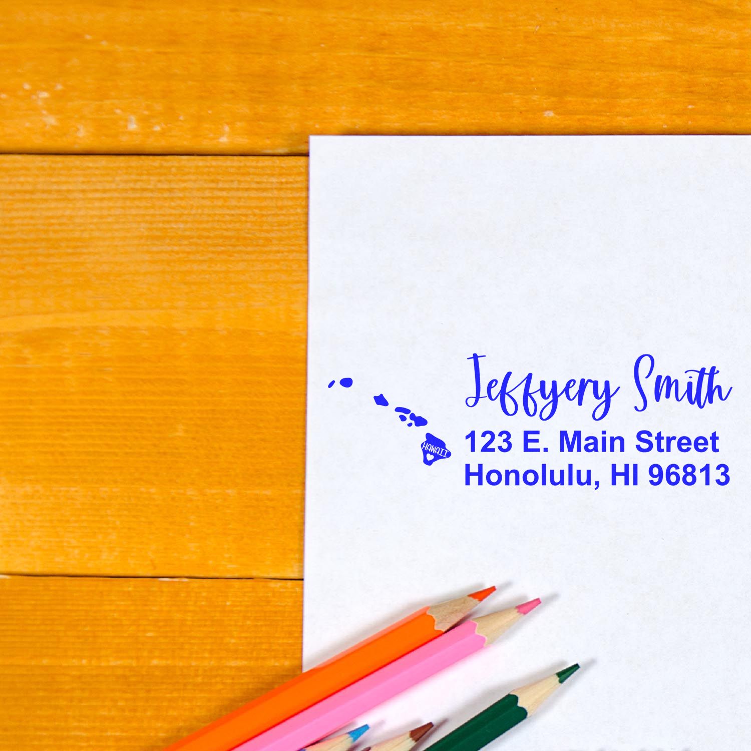 Slim Hawaii Custom Address Stamp for Envelopes on white paper with blue ink, featuring a map of Hawaii. Colored pencils are nearby on a wooden surface.