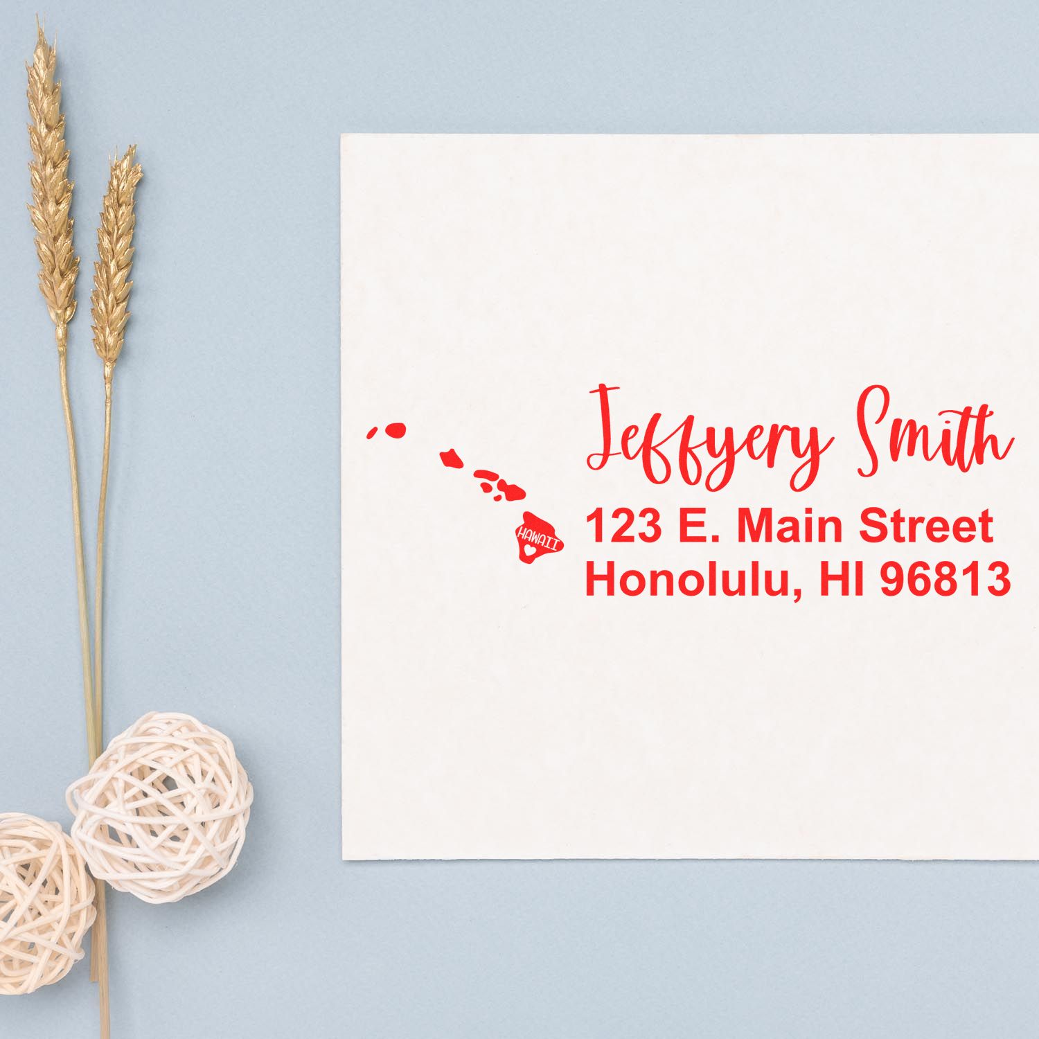 Slim Hawaii Custom Address Stamp for Envelopes in red ink on a white card, featuring a map of Hawaii. The card is placed on a light blue surface with decorative elements nearby.