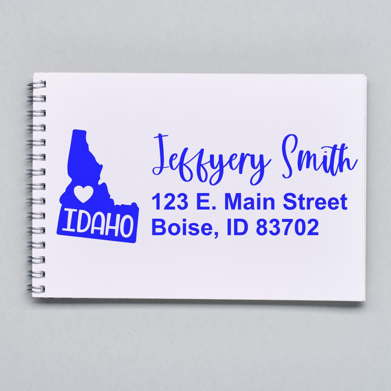 Image of a notebook displaying the Slim Idaho Custom Address Stamp for Envelopes, featuring a blue Idaho state outline with a heart and sample address text in a stylish font.