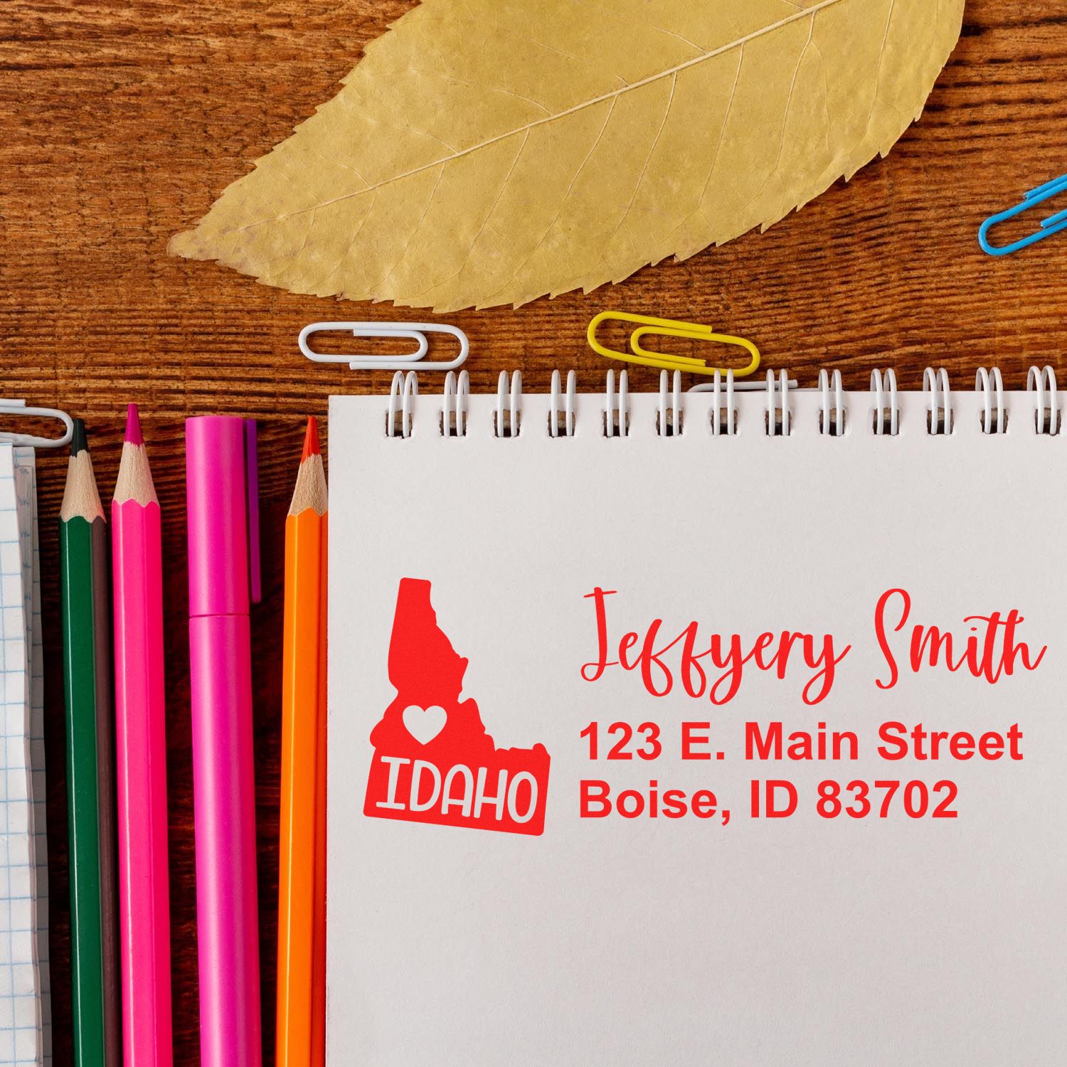 Idaho State Love Personalized Address Stamp on a notepad with colored pencils and paperclips, featuring a red Idaho map design and address text in red ink on a wooden surface.