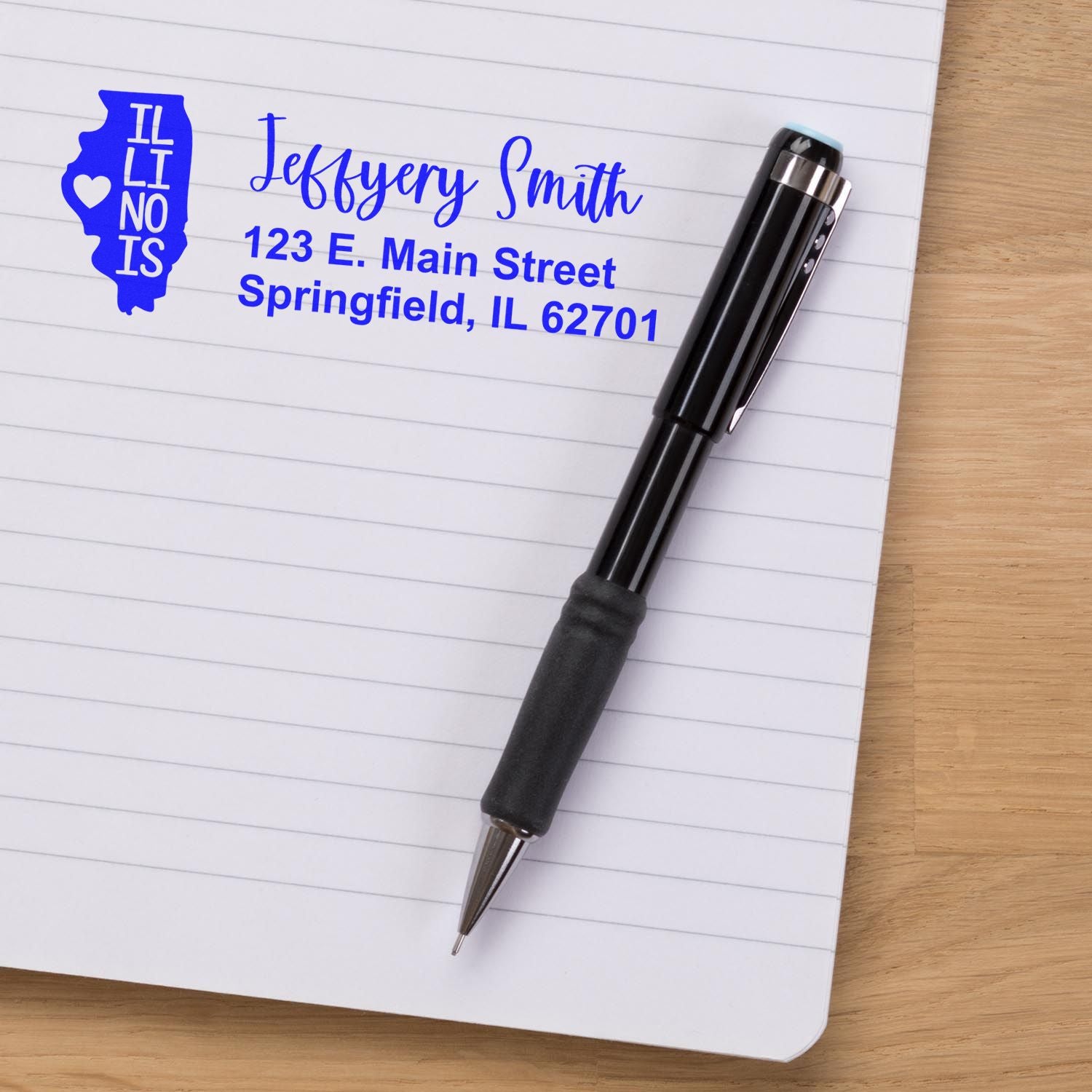 Slim Illinois Custom Address Stamp for Envelopes displayed on a notebook page with a pen. The stamp shows a sample address in blue ink, featuring an Illinois state outline design.