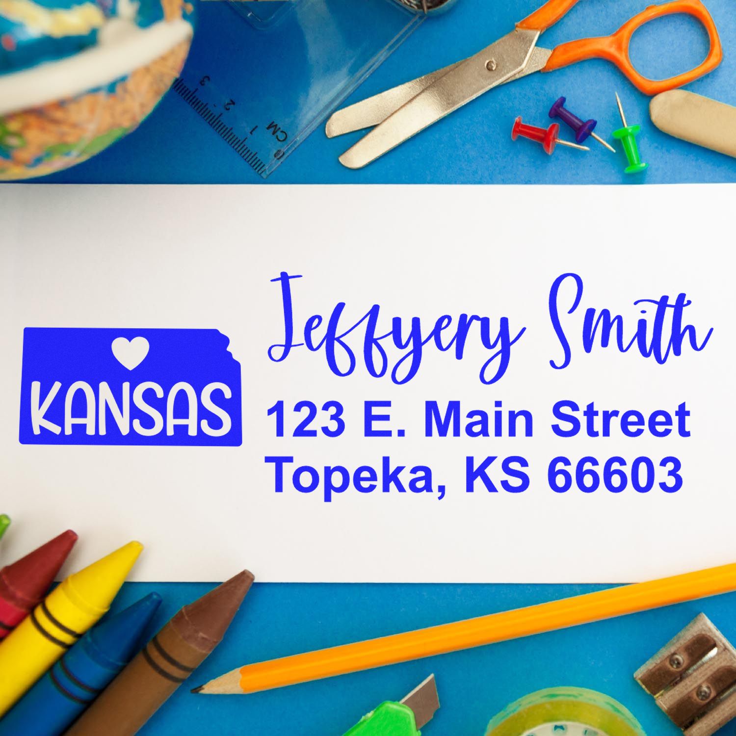 Slim Kansas Custom Address Stamp for Envelopes on a white envelope with colorful stationery items like scissors, pins, and crayons around it. The stamp features a heart and the word Kansas.