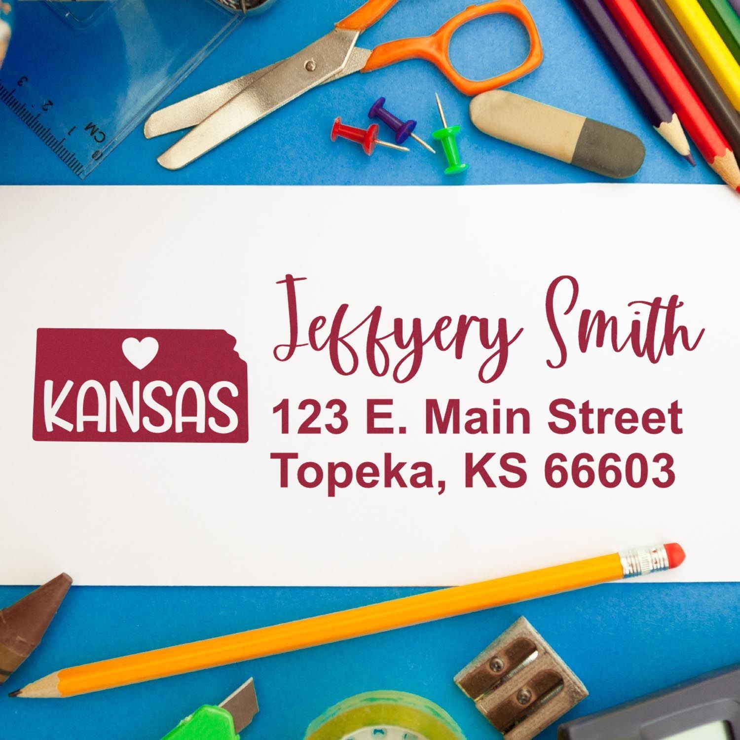 PSI Pre-Inked Kansas State Love Customized Address Stamp on white paper with Jeffery Smith, 123 E. Main Street, Topeka, KS 66603 in red. Surrounded by colorful office supplies on a blue surface.