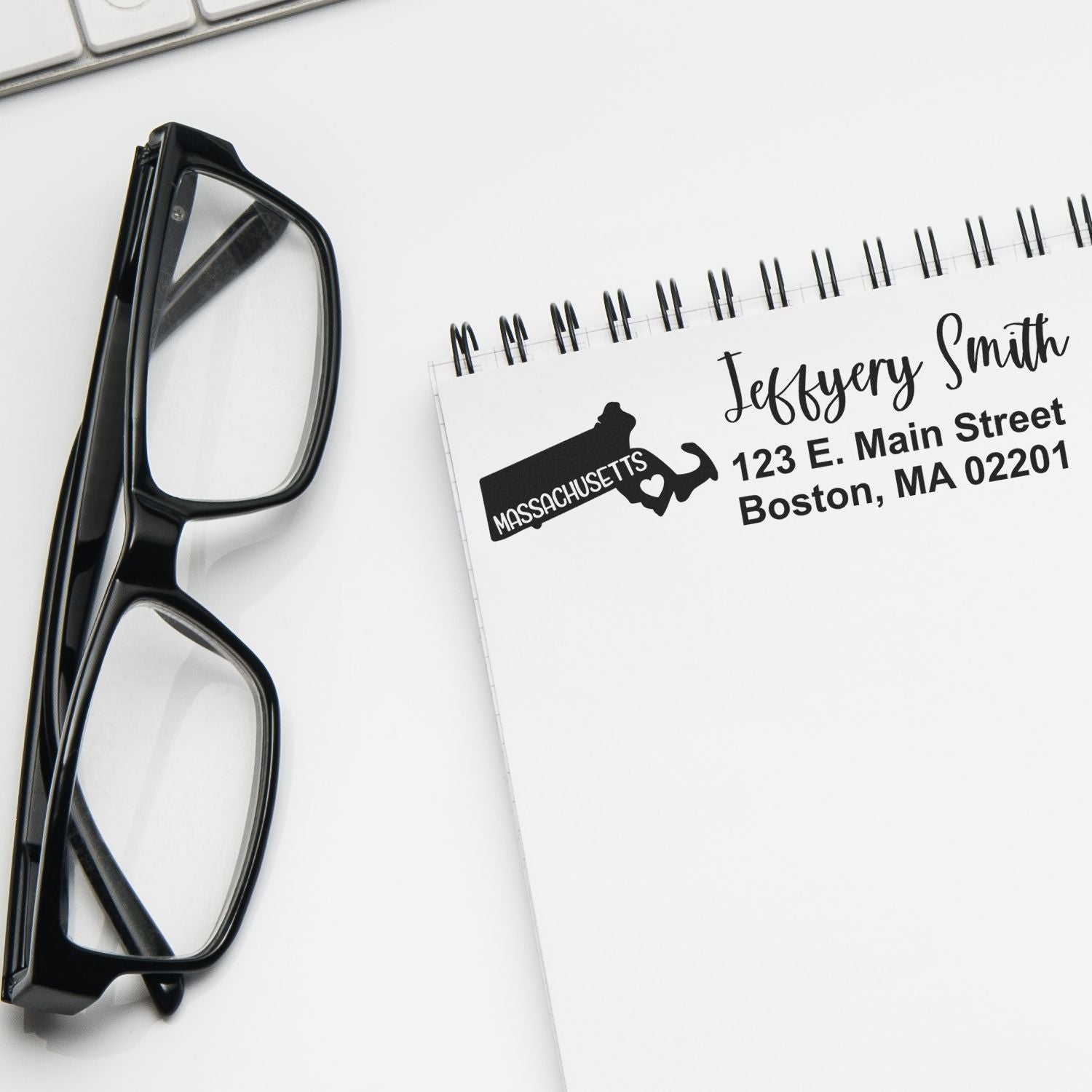 Slim Massachusetts Custom Address Stamp for Envelopes displayed on a notepad with a pair of glasses nearby. The stamp shows a sample address in a stylish font, ideal for personalizing mail.