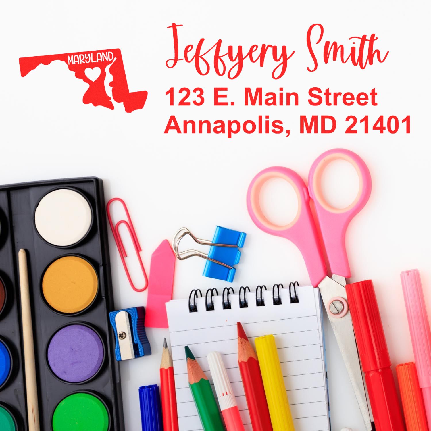 A colorful desk with art supplies and a notepad featuring the State Love of Maryland Custom Address Stamp Self-Inking, displaying Jeffery Smith, 123 E. Main Street, Annapolis, MD 21401 in red.