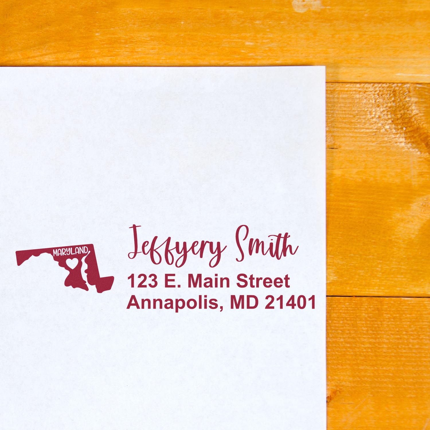 Maryland State Love Personalized Address Stamp on white envelope with red ink, featuring a heart design and address: 123 E. Main Street, Annapolis, MD 21401, on a wooden surface.