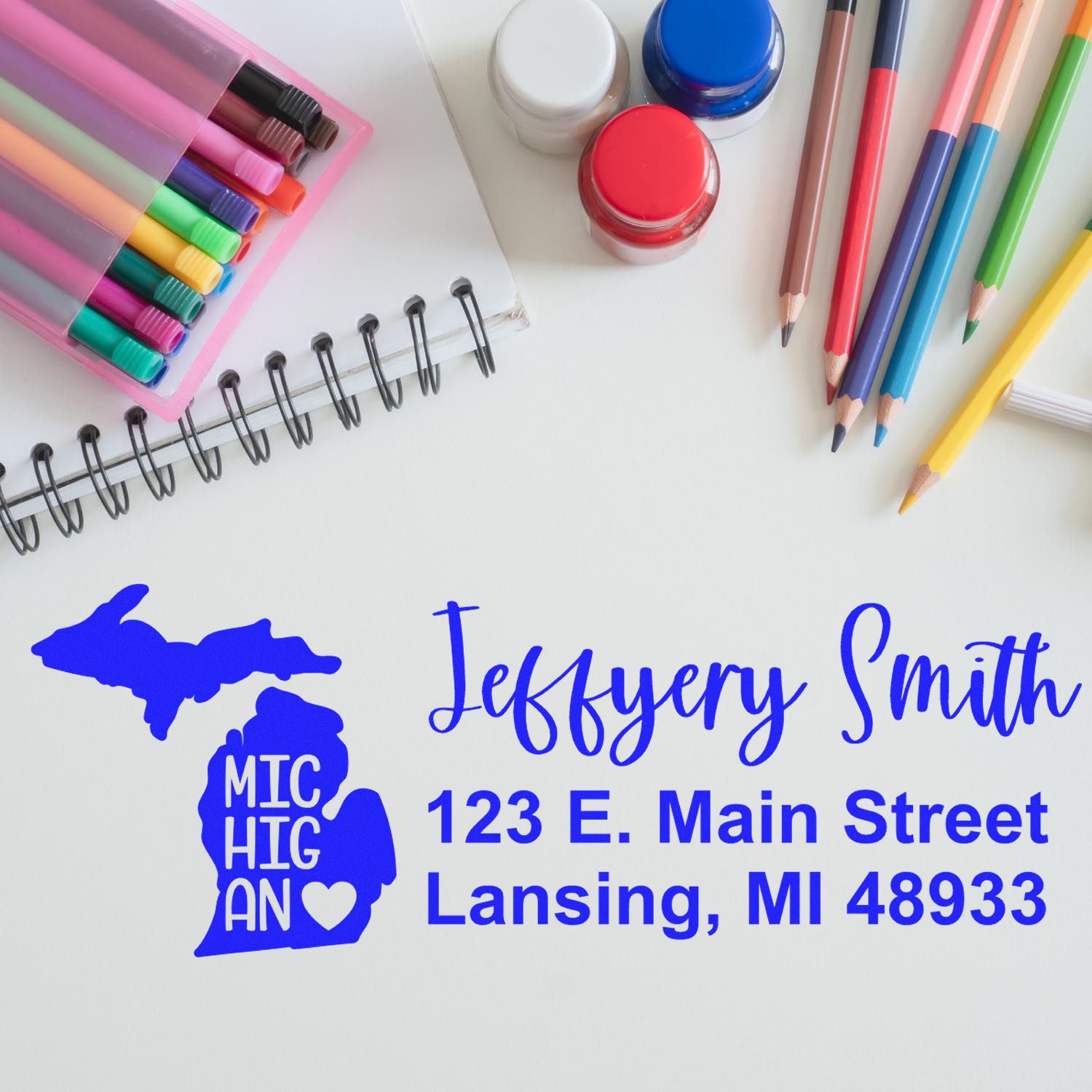 Desk with colorful pens, notebook, and PSI Pre-Inked Michigan State Love Customized Address Stamp showing Jeffery Smith, 123 E. Main Street, Lansing, MI 48933 with Michigan map design.