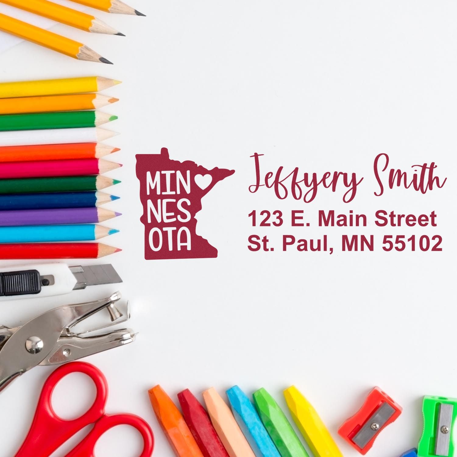 Colorful pencils, scissors, and a hole punch surround an envelope stamped with the Slim Minnesota Custom Address Stamp, featuring a red Minnesota state outline and personalized address text.