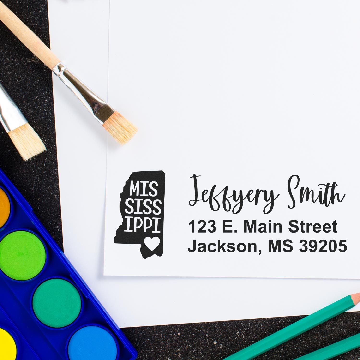 State Love of Mississippi Custom Address Stamp Self-Inking on paper with paintbrushes and watercolor palette, featuring a Mississippi state outline and personalized address.