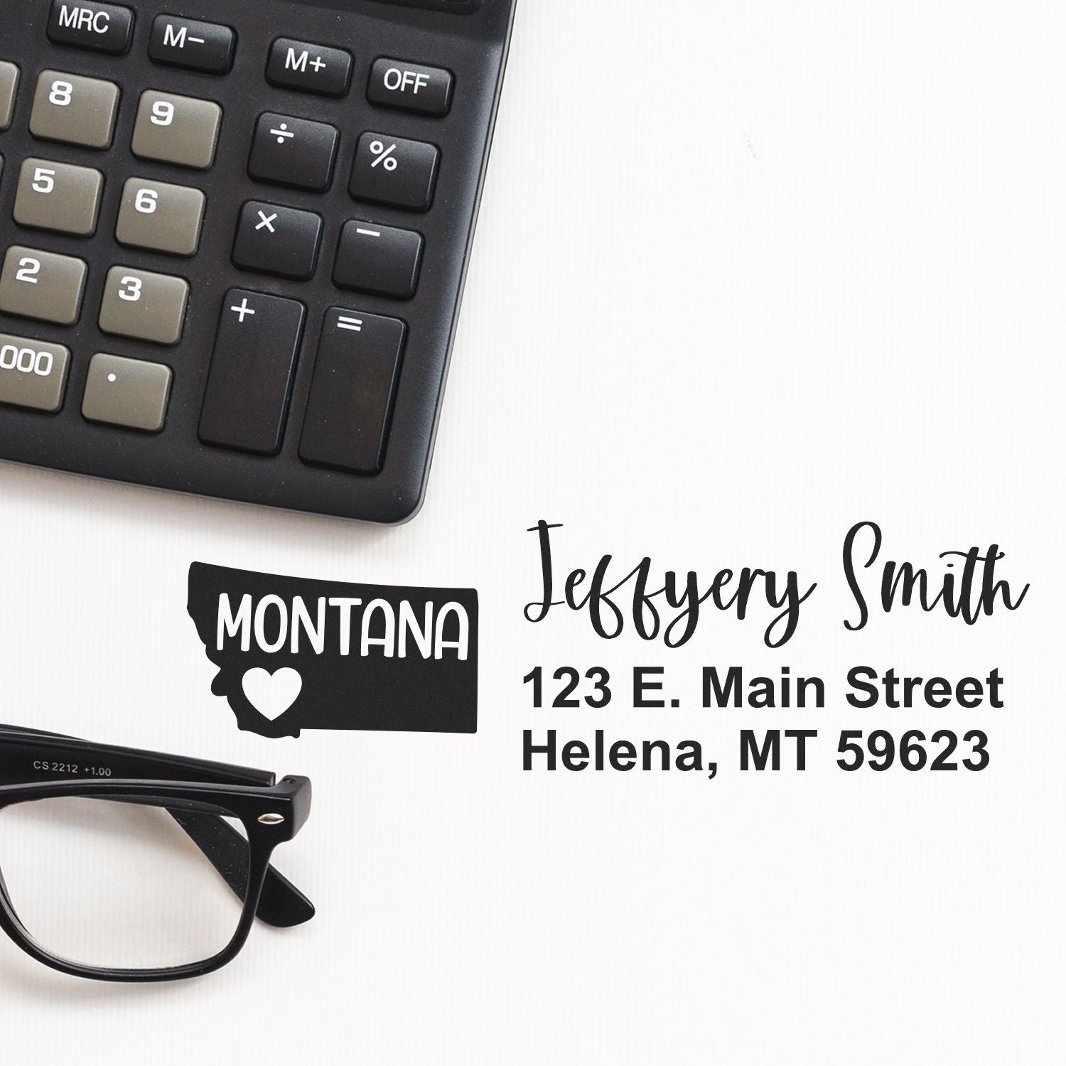 State Love of Montana Custom Address Stamp Self-Inking displayed on a desk with a calculator and glasses, featuring a heart within the Montana state outline and personalized address details.