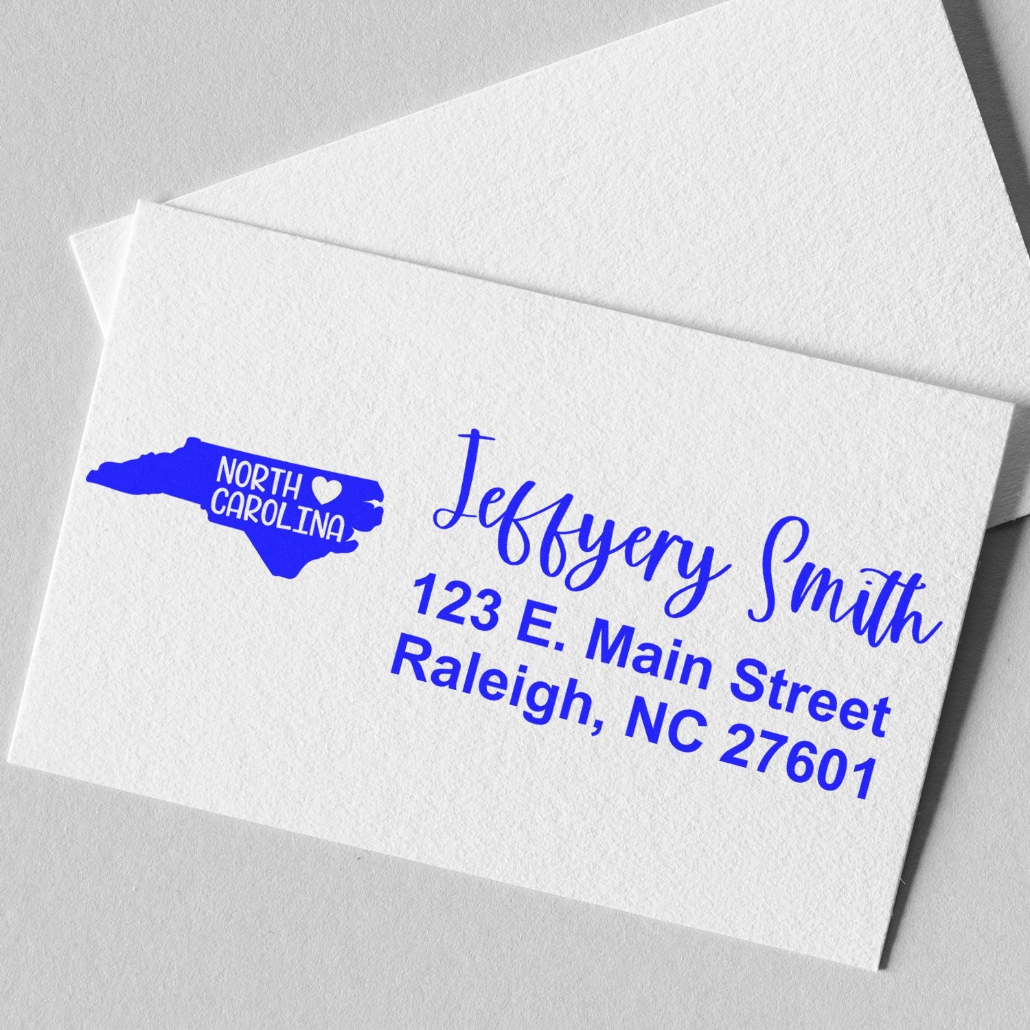 North Carolina State Love Personalized Address Stamp on a white card, featuring a blue state outline with a heart and bold, elegant text displaying a name and address in Raleigh, NC.