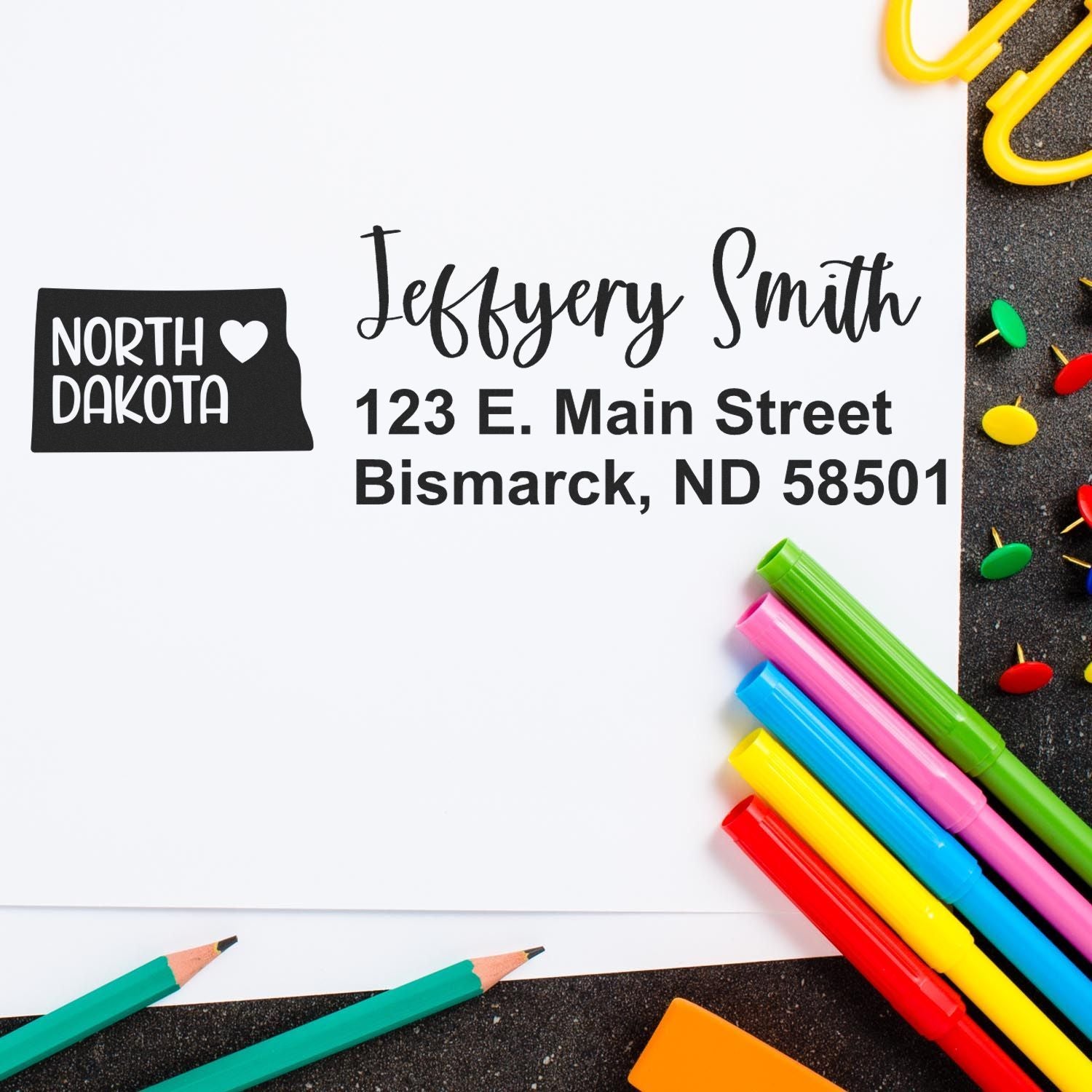 North Dakota State Love Personalized Address Stamp on white paper with colorful pens and paperclips. The stamp features a heart and state outline, showcasing an address in Bismarck, ND.