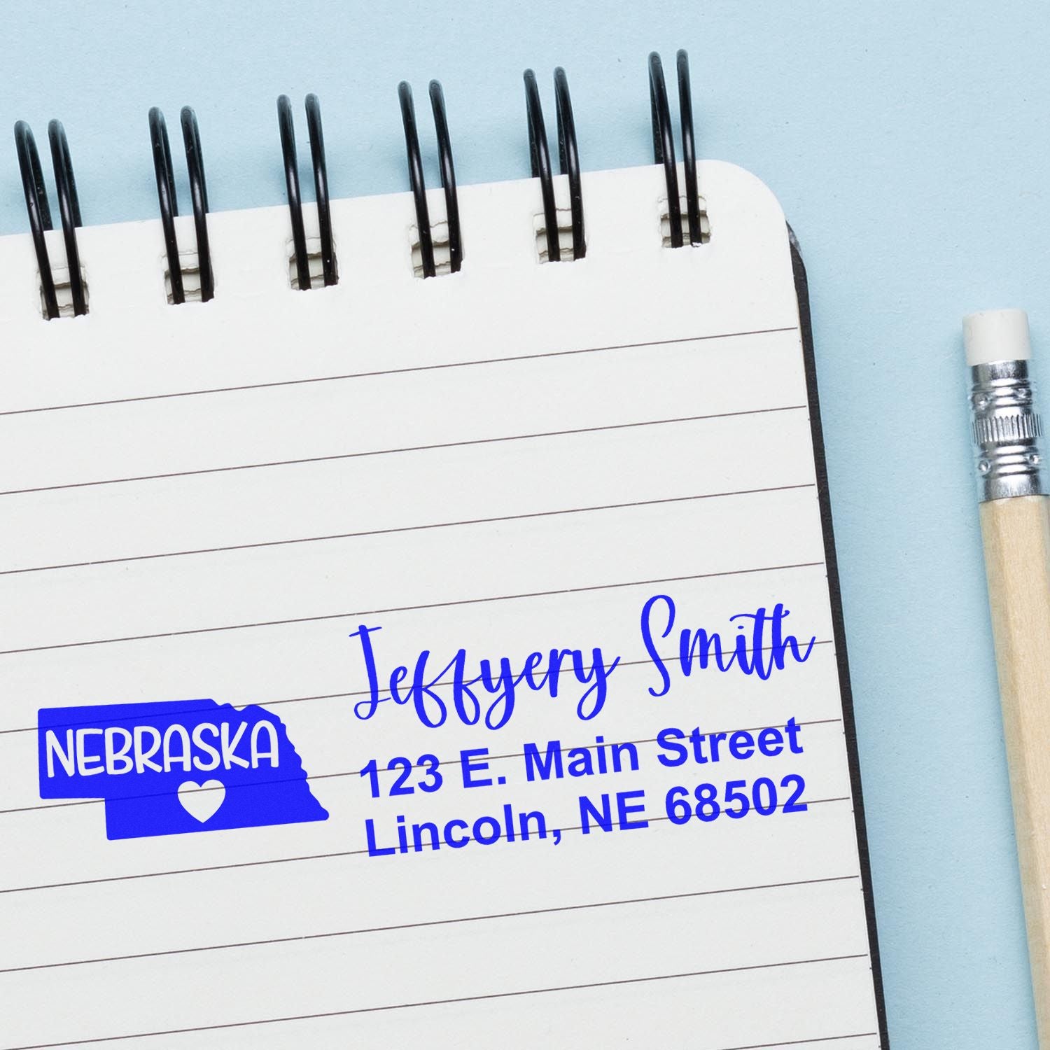 PSI Pre-Inked Nebraska State Love Customized Address Stamp on a notebook page with a pencil, displaying a sample address in blue ink featuring a Nebraska state outline with a heart.