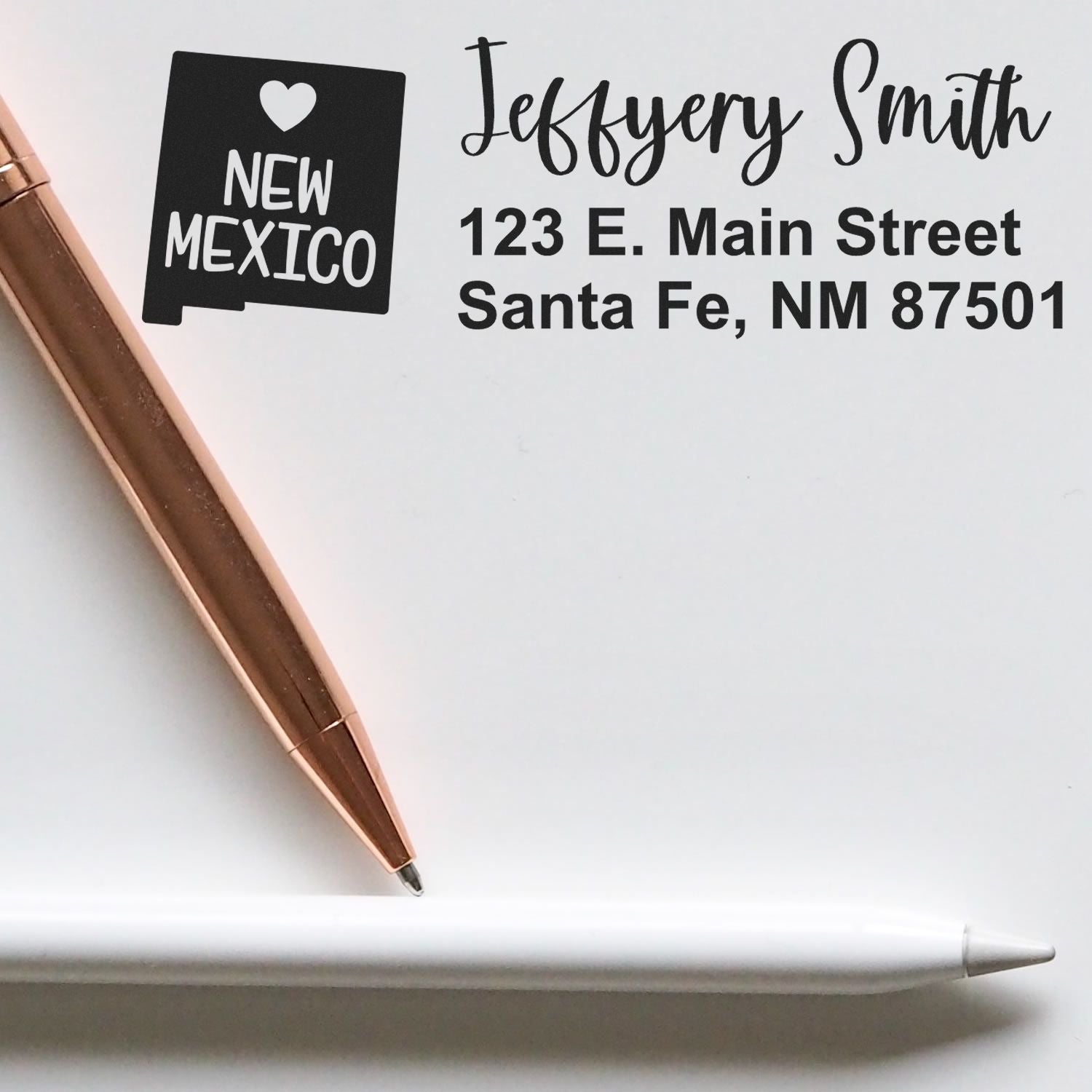 PSI Pre-Inked New Mexico State Love Customized Address Stamp on paper with 'Jeffery Smith, 123 E. Main Street, Santa Fe, NM 87501' next to a rose gold pen and a white stylus.