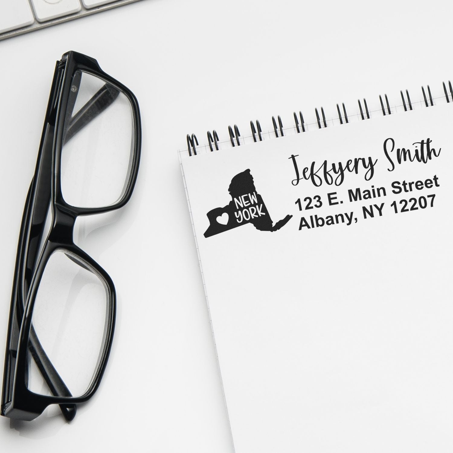 Slim New York Custom Address Stamp for Envelopes displayed on a notepad with a New York state outline and address. Glasses and a keyboard are nearby, highlighting its use for personalizing mail.